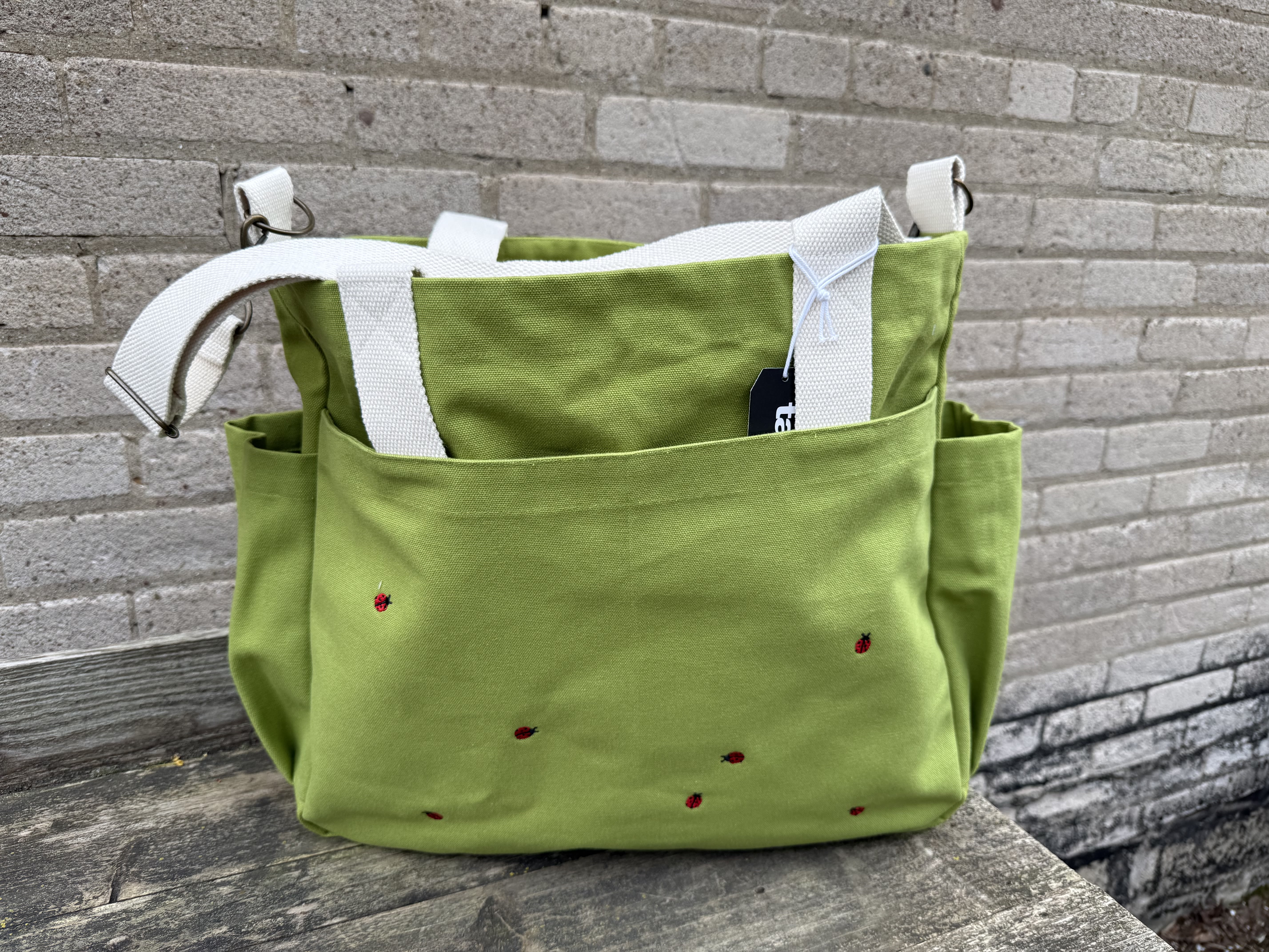 Bee Garden Carry Bag - A bright green canvas tote with playful red ladybug embroidery - the Bee Garden Carry Bag makes a cheerful everyday gift for birthdays, Mother's Day, or just because you're thinking of her. As a practical carry-all, this tote conveys a light, joyful message: she'll feel seen and smiled at every time she reaches for it, whether headed to market, the garden, or a friend's house.  The bag's structured, boxy shape and roomy interior are balanced by wide white webbing straps and sturdy metal rings for reliable wear. Side pockets add convenient organization, while a hanging tag gives a finished, gift-ready look. The green canvas paired with white straps and small red embroidered ladybugs creates a fresh, springlike palette that pairs easily with a hand-tied posy or potted plant.  Perfect as a container for florals or as a standalone gift, this tote suits recipients who appreciate practical, whimsical design and outdoor-loving style. Order with same-day pickup or delivery when available; wipe clean canvas and avoid heavy soaking to keep the colors bright.