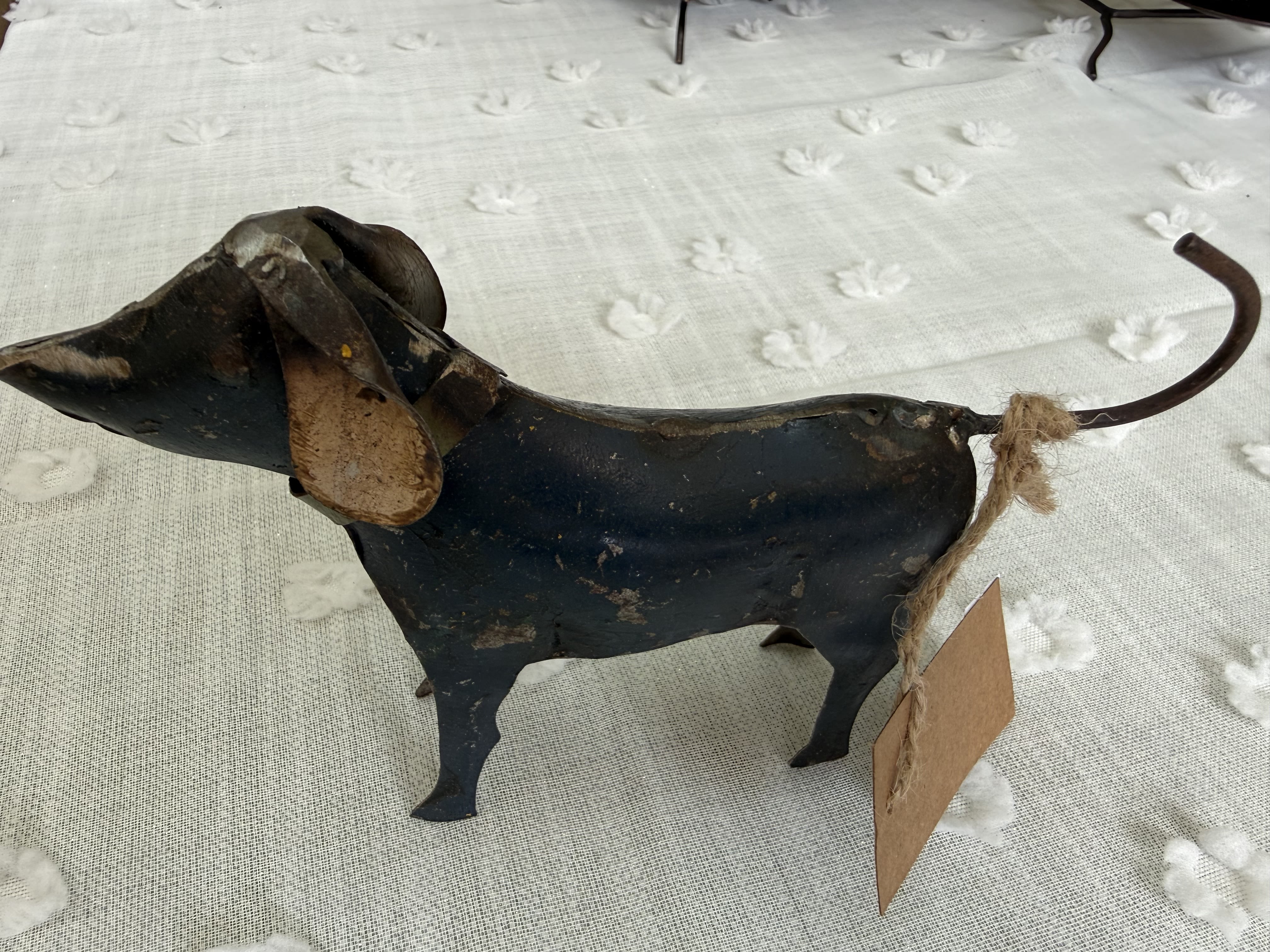 Cute Metal Dog - A whimsical metal dog figurine brings rustic charm to any gift moment-perfect as a small flower arrangement accent or a standalone keepsake for Mother's Day, Father's Day, birthdays, anniversaries, or "just because" surprises. The playful standing pose and curled tail create an immediate, heartwarming impression that will make the person you're thinking of feel noticed and adored, whether you're celebrating a new baby, a graduation, or saying "I'm sorry."  Hand-forged in dark metal with a worn, vintage finish, this petite figure has a rope-tied tail and a paper tag that add handmade character and tactile contrast to bouquets or tabletop vignettes. It looks striking against light linens or nestled among black-and-white blooms and seasonal greens; use it as a lawn-side token for Memorial Day or a cozy mantelpiece accent through Thanksgiving and Christmas. Sized to pair easily with compact arrangements, this durable keepsake works year-round and requires no care-just place where it can be seen and enjoyed.