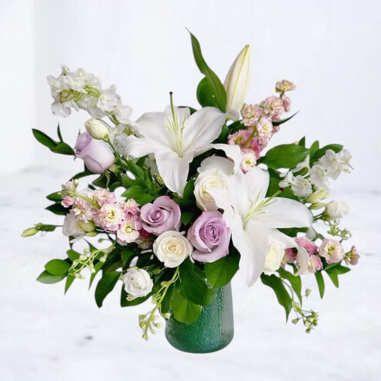Lavender Solace Bouquet - The Lavender Solace Bouquet offers a peaceful embrace through soft hues of ivory, blush, and lavender. Featuring elegant white lilies, delicate lavender roses, white spray roses, and stock, this arrangement brings comfort and beauty to sympathy occasions and beyond. Hand-arranged in a soft green glass vase, it's a gentle and serene gift for memorial services, condolence deliveries, or thoughtful gestures of remembrance.  Available for same-day delivery in League City, Dickinson, Galveston, and surrounding areas.  Substitution Policy: At Twigs and Blooms, we craft every bouquet using the freshest blooms in season. Should substitutions be necessary, we will maintain the overall integrity, style, and color palette of the arrangement.