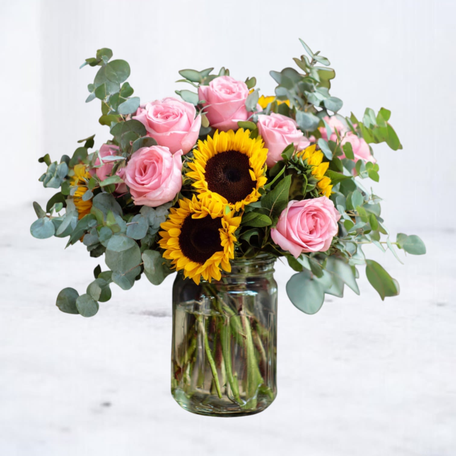 Medium bouquet of pink roses and bright sunflowers in a clear glass jar vase with eucalyptus.