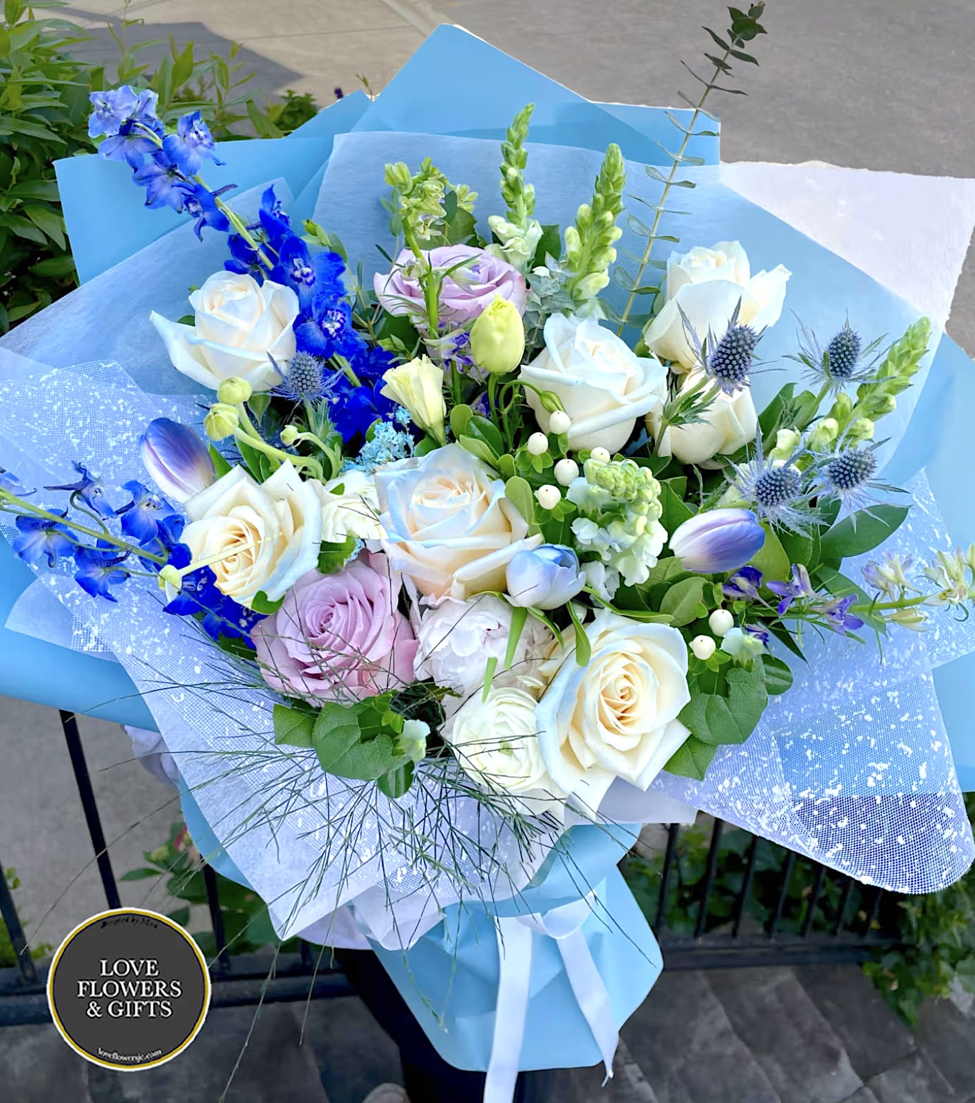 Hand-tied bouquet of white roses, blue delphinium, and pastel blooms wrapped in blue paper.