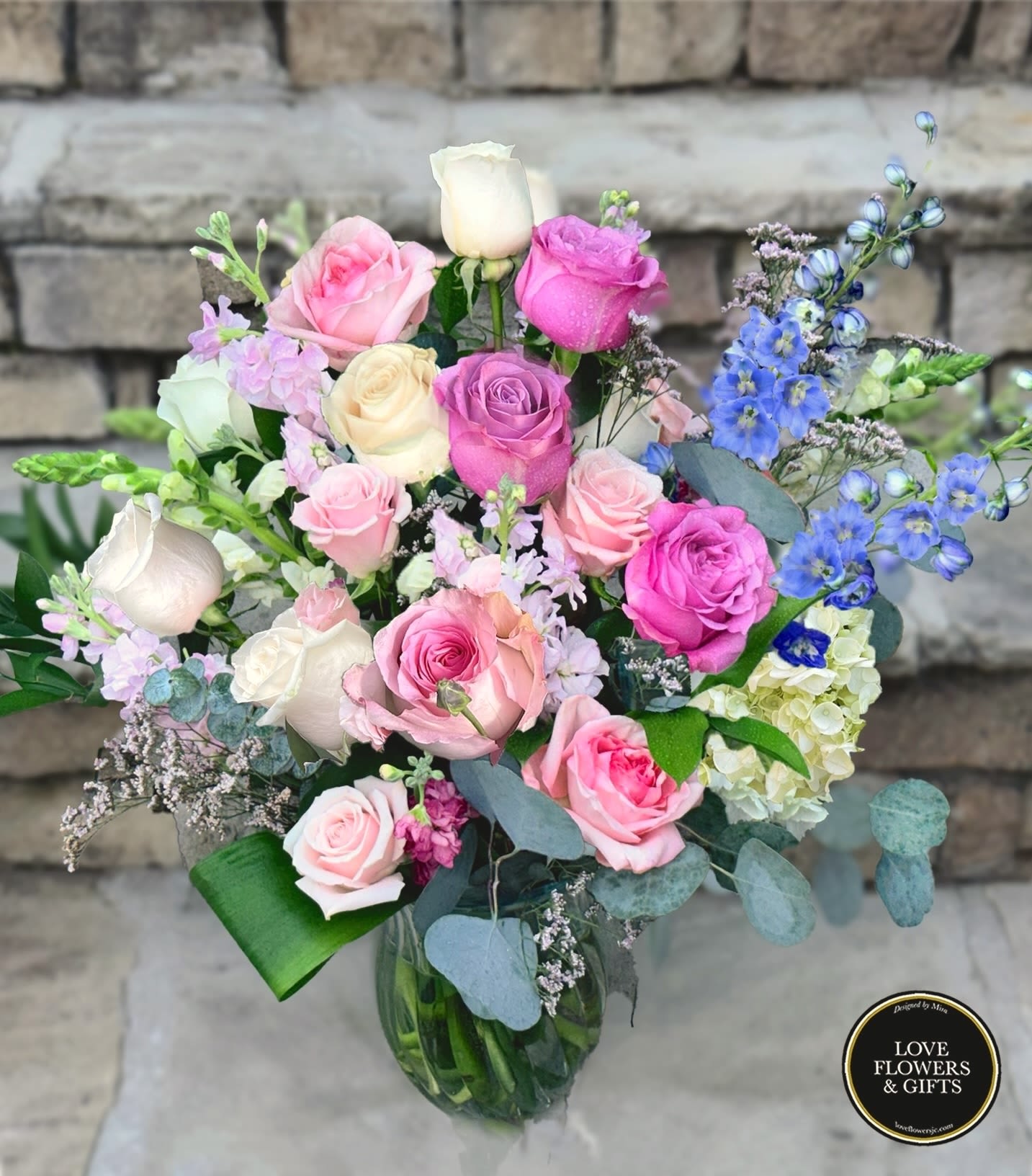 Mixed bouquet of pink and white roses with blue delphinium in a clear glass vase.