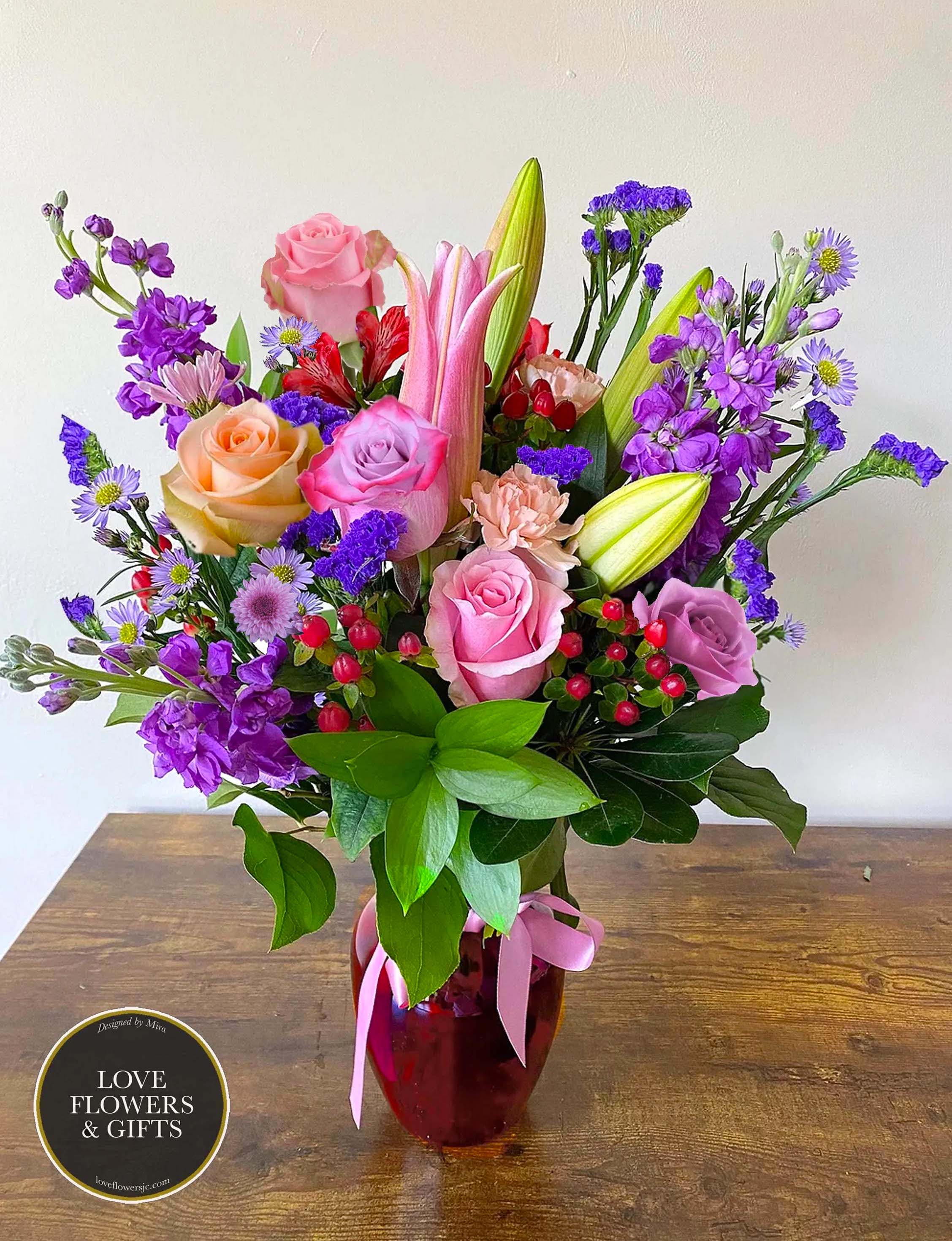 Mixed bouquet of pink roses, lilies, and purple flowers in a red glass vase with pink ribbon.