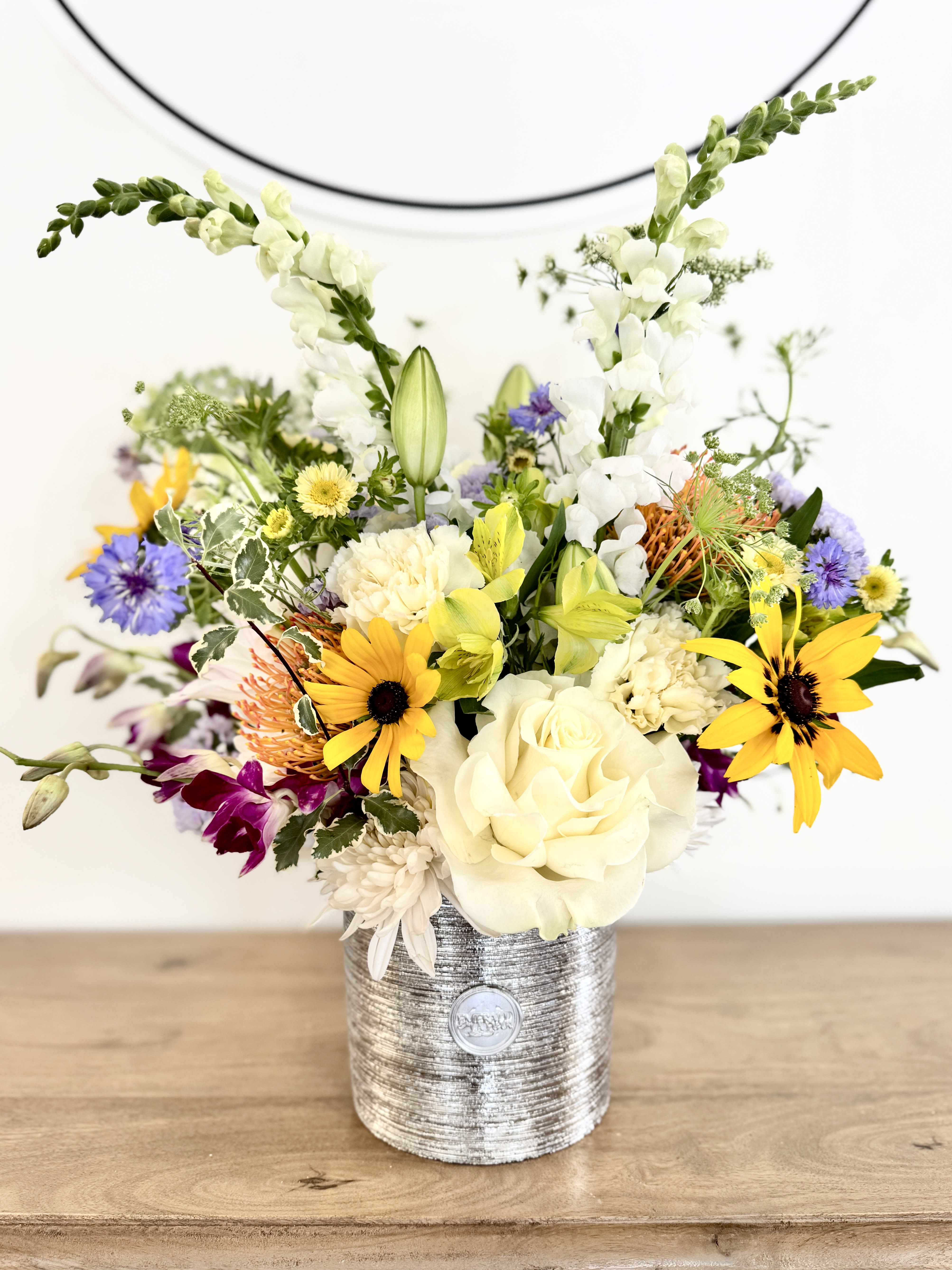 Silver Strength - Designed to honor the men who lead with both strength and heart, Silver Strength is a sophisticated floral statement arranged in a striking silver vessel. Thoughtfully curated with premium seasonal blooms, textural elements, and architectural lines, this design balances masculine presence with an unexpected softness.  Featuring a refined blend of distinctive florals and natural movement, Silver Strength is created for fathers, mentors, husbands, grandfathers, and the remarkable men who deserve recognition. Its elevated scale and artistic composition make it a meaningful gesture that feels both substantial and heartfelt.  Perfect for Father's Day, milestone celebrations, professional achievements, or simply expressing gratitude, Silver Strength offers a modern interpretation of floral gifting for him. Confident, sophisticated, and memorable, it is designed to leave a lasting impression while celebrating the strength found in kindness, wisdom, and grace.