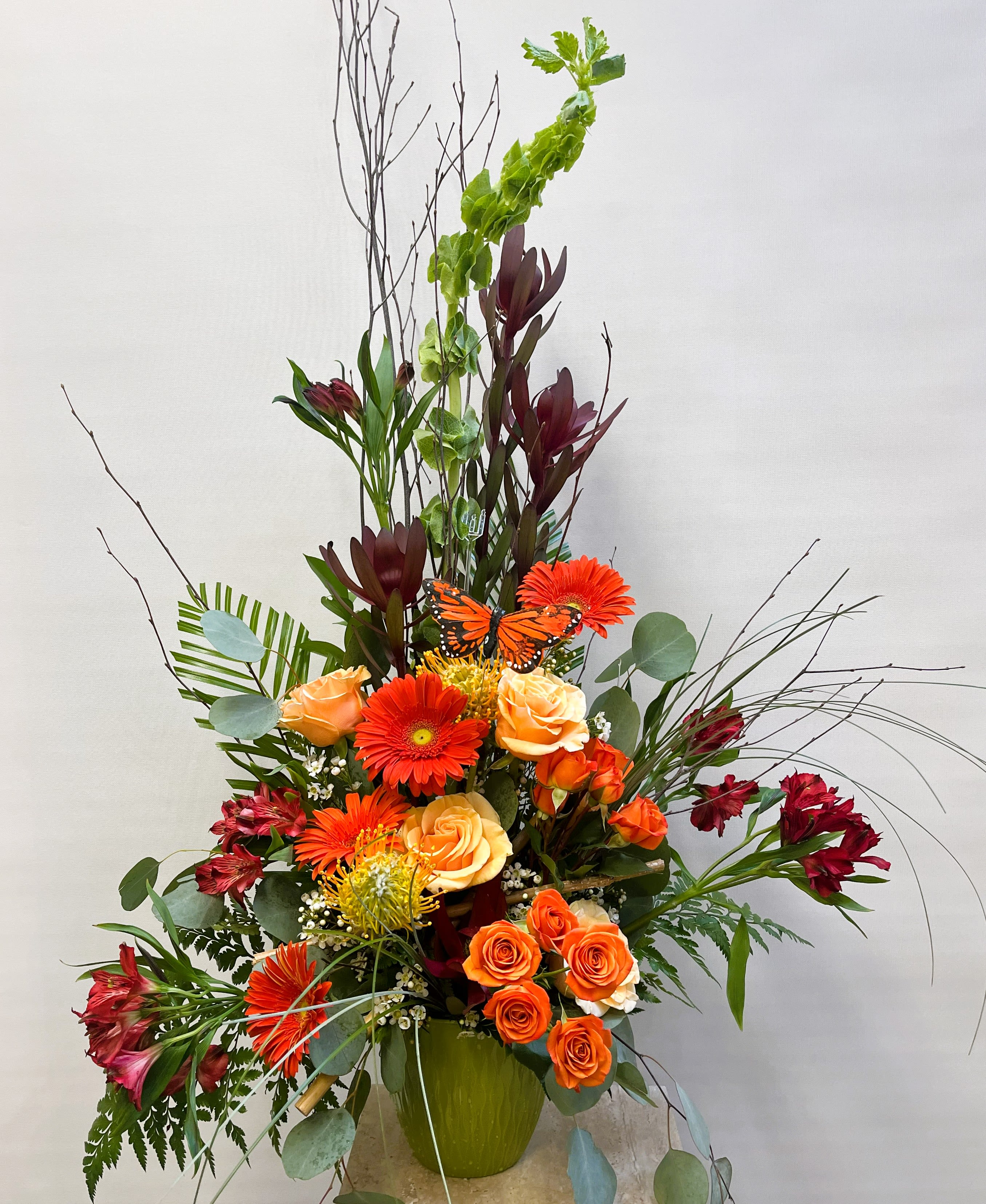 Tall floral arrangement with orange roses, gerbera daisies, and burgundy blooms in a green vase