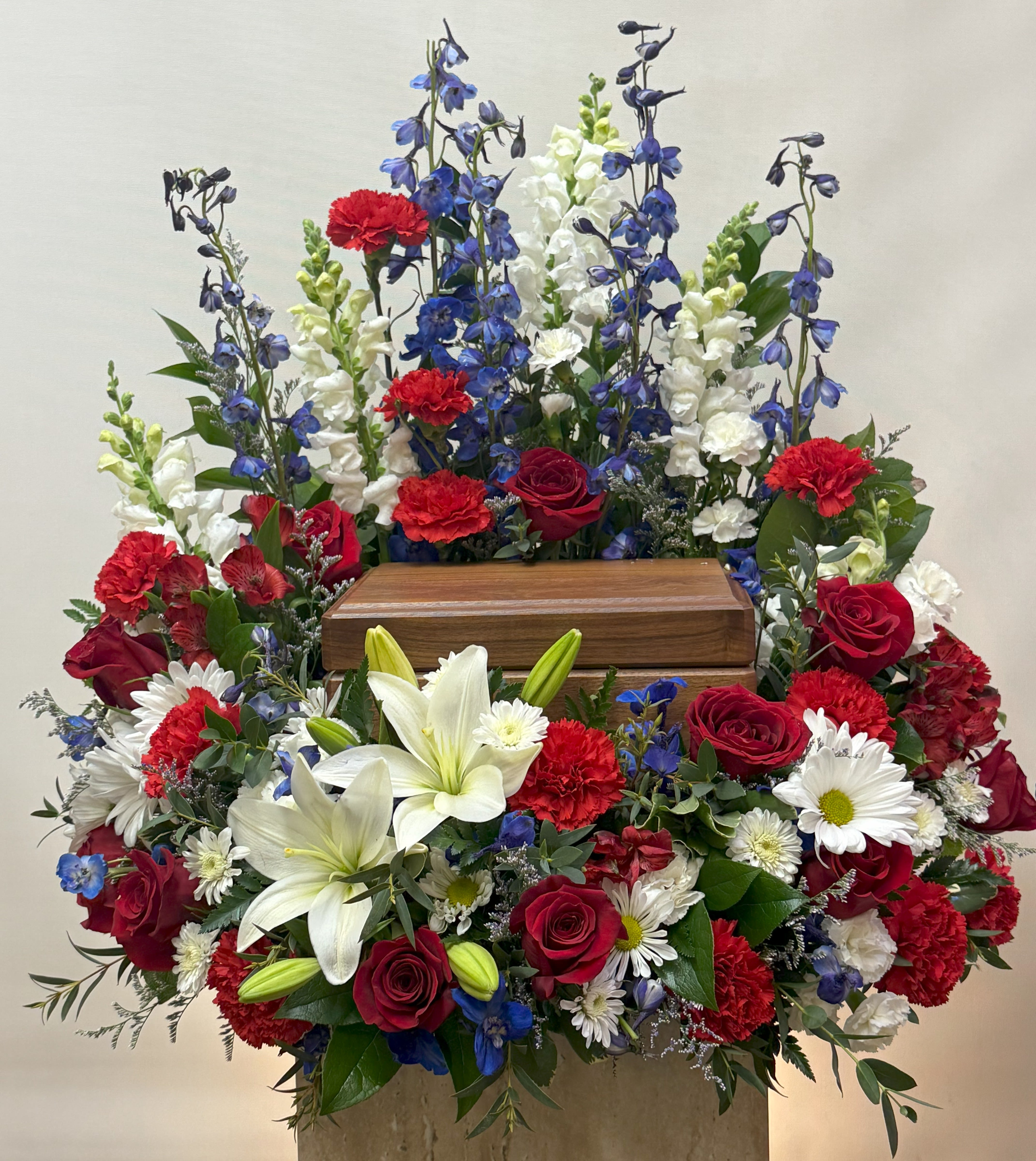 Large funeral spray with red roses, white lilies, and blue delphinium around a wooden urn