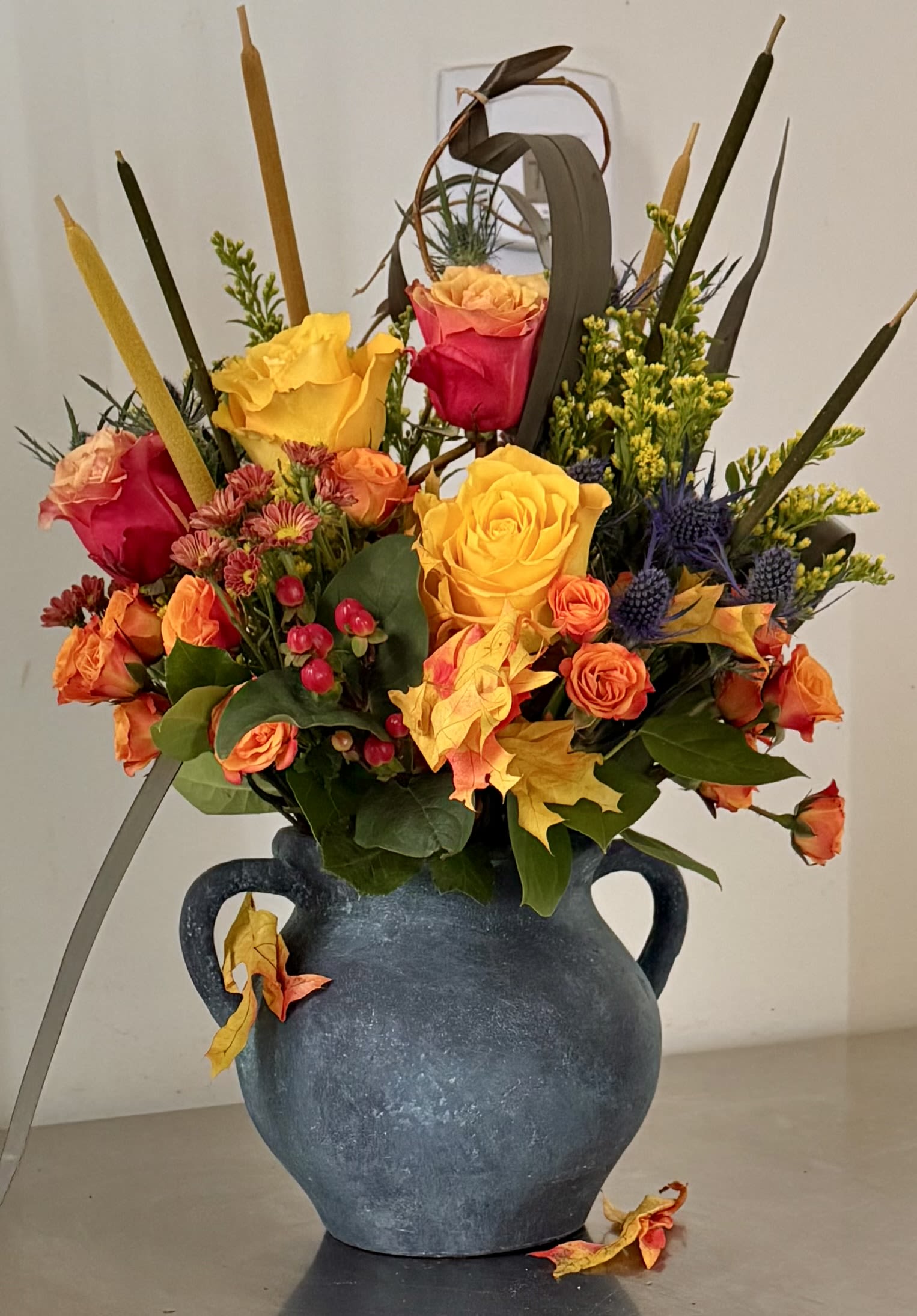 Autumn Grandeur  - A premium, large, autumn-themed design with a wildflower look capturing the essence of the autumn season.  Features a vibrant mix of yellow, orange, roses, hypericum berries, thistle, and various autumn leaves and greenery. Presented in a unique, handmade clay vase with patina. 