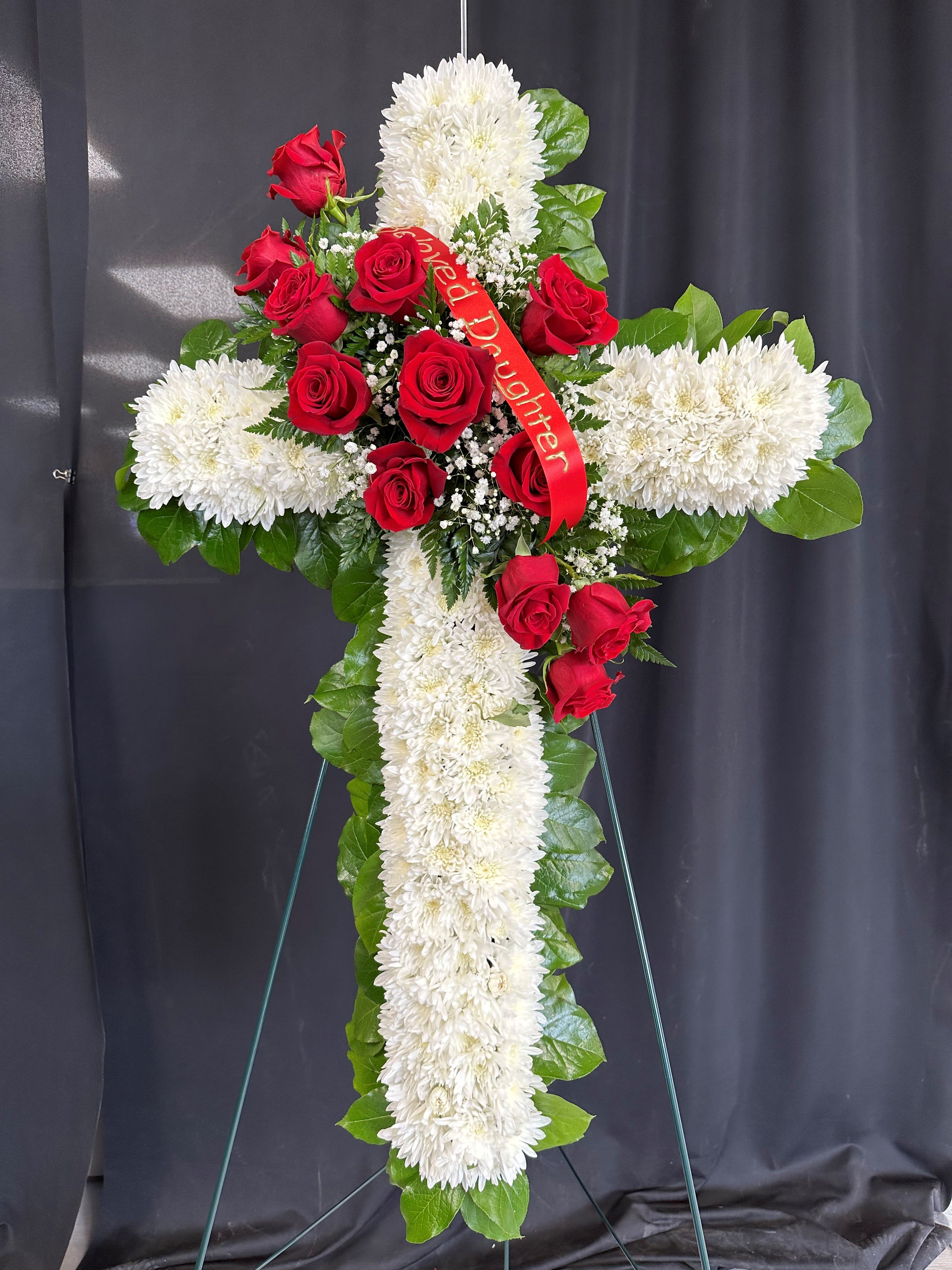 Tall white chrysanthemum cross with red roses and a red memorial ribbon on a metal easel.