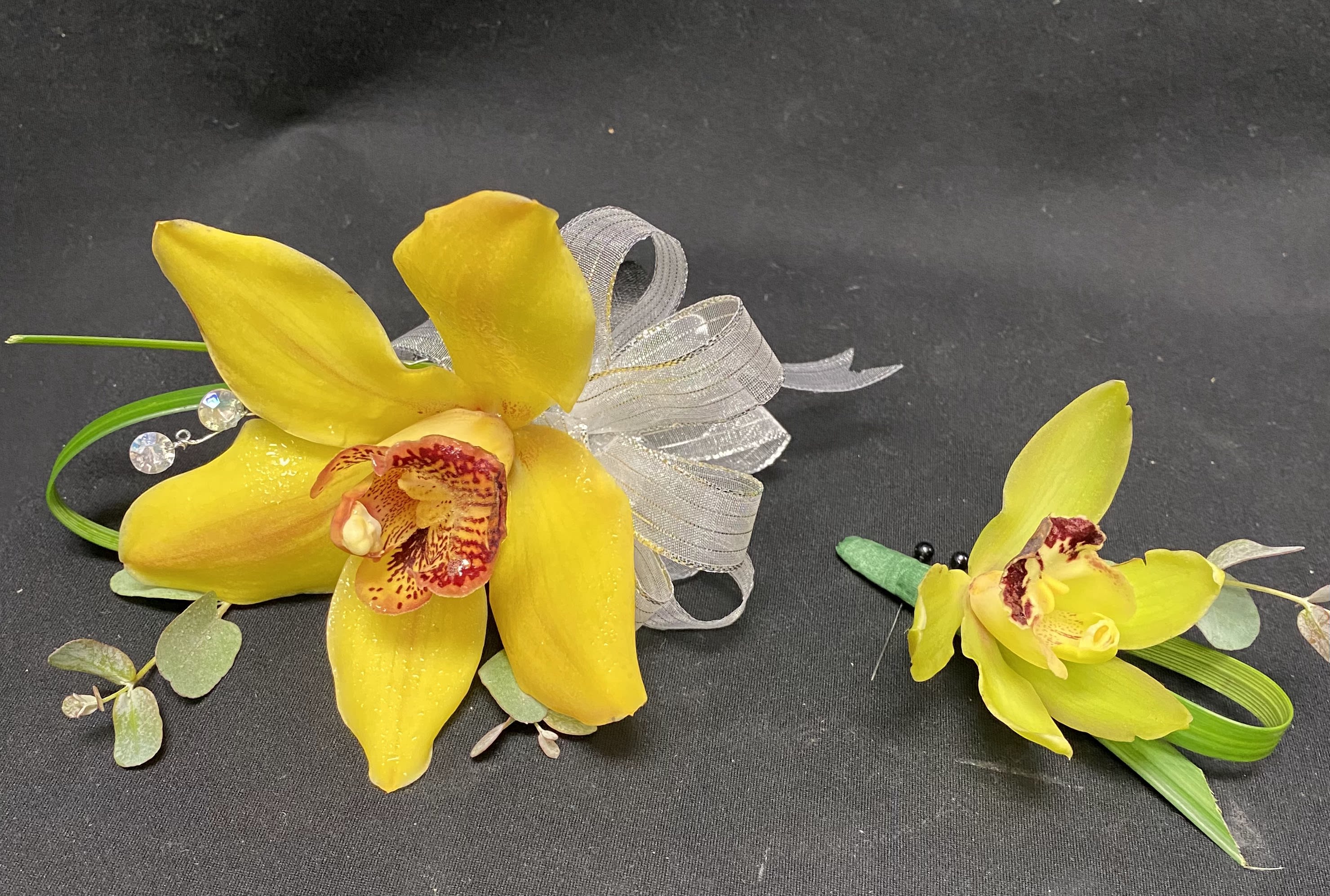 Two yellow orchid corsages with ribbon and greenery on a dark background