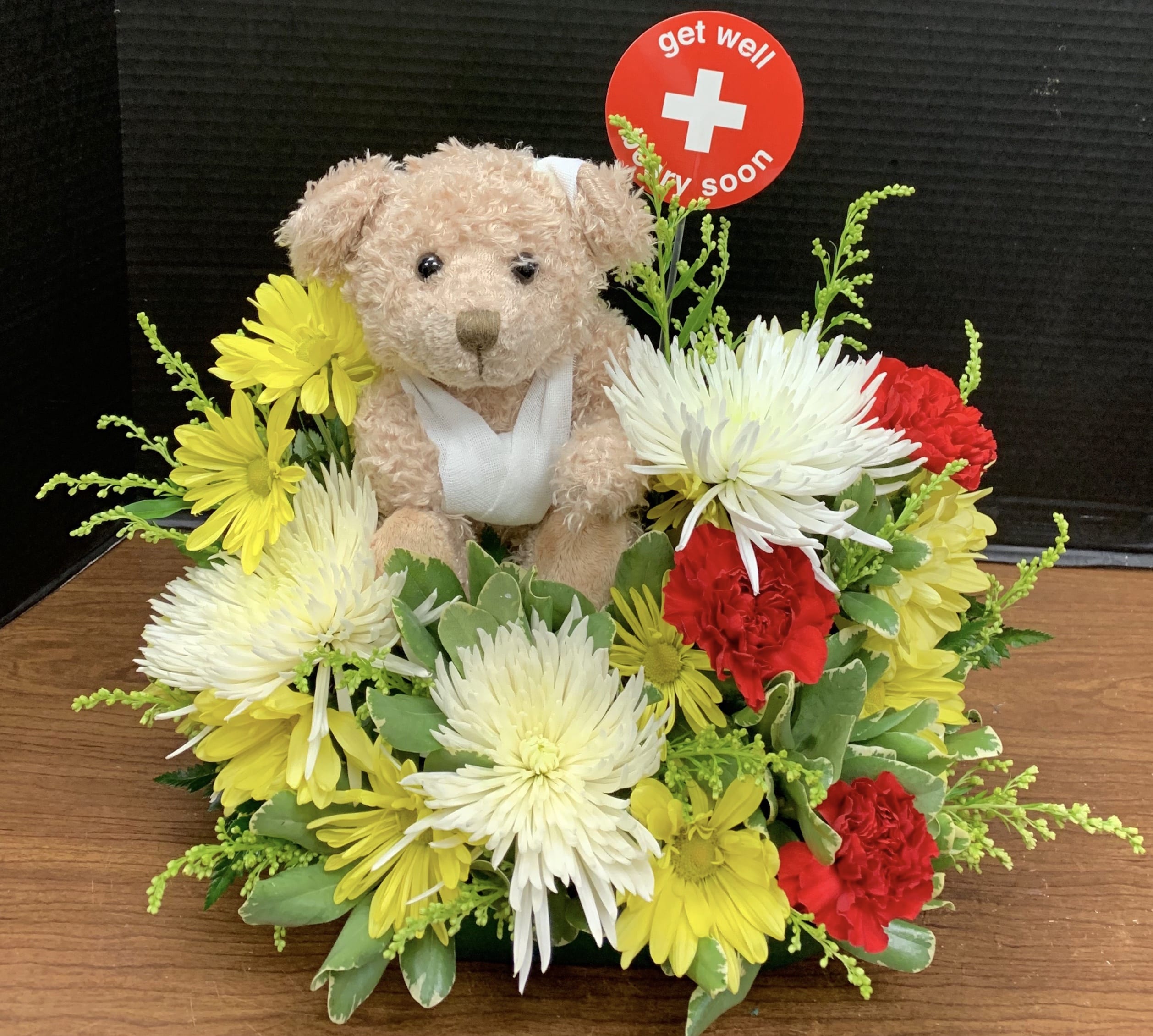 Teddy bear surrounded by yellow, white, and red flowers with a get well sign