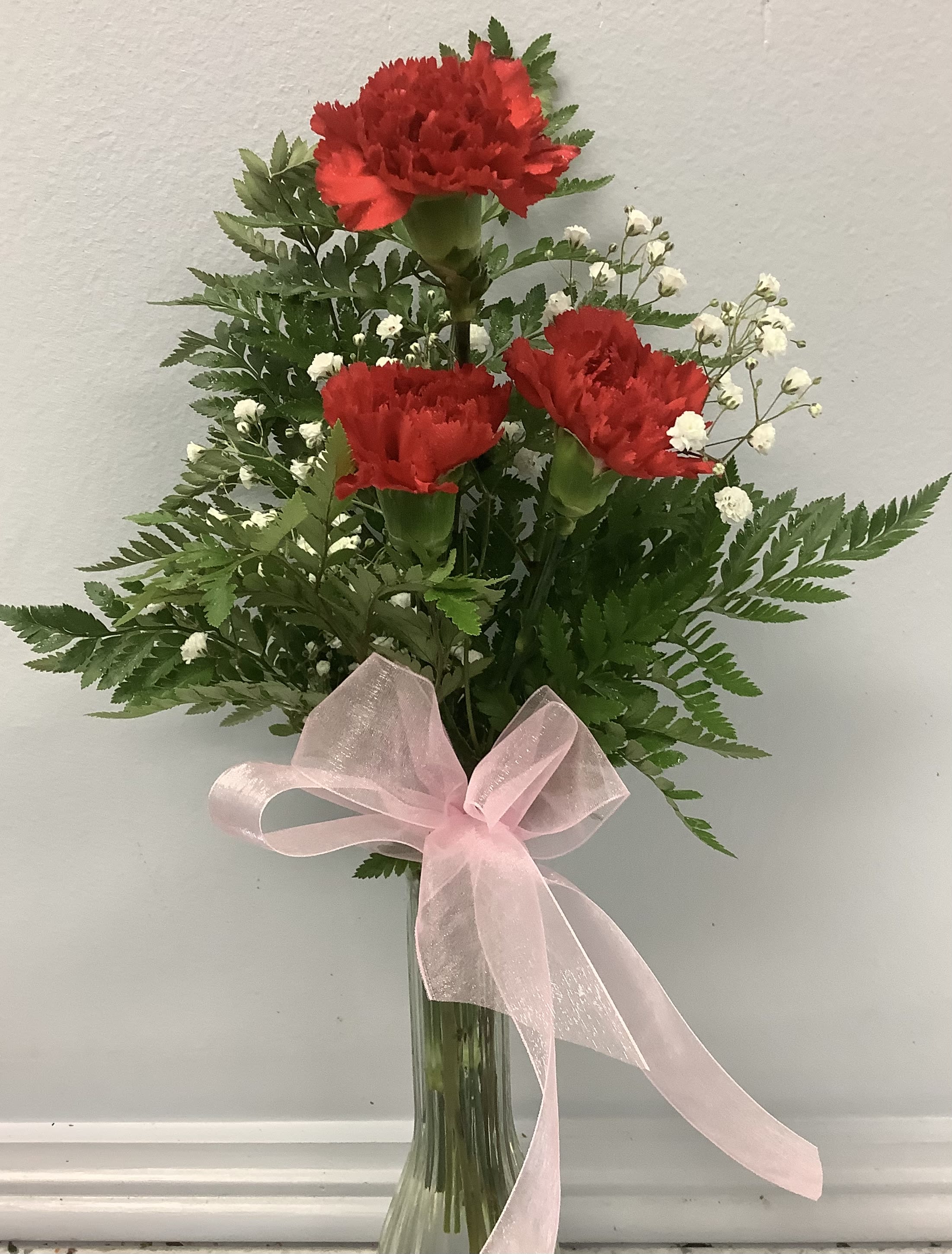 Red carnations in a glass vase with baby’s breath and a pink ribbon