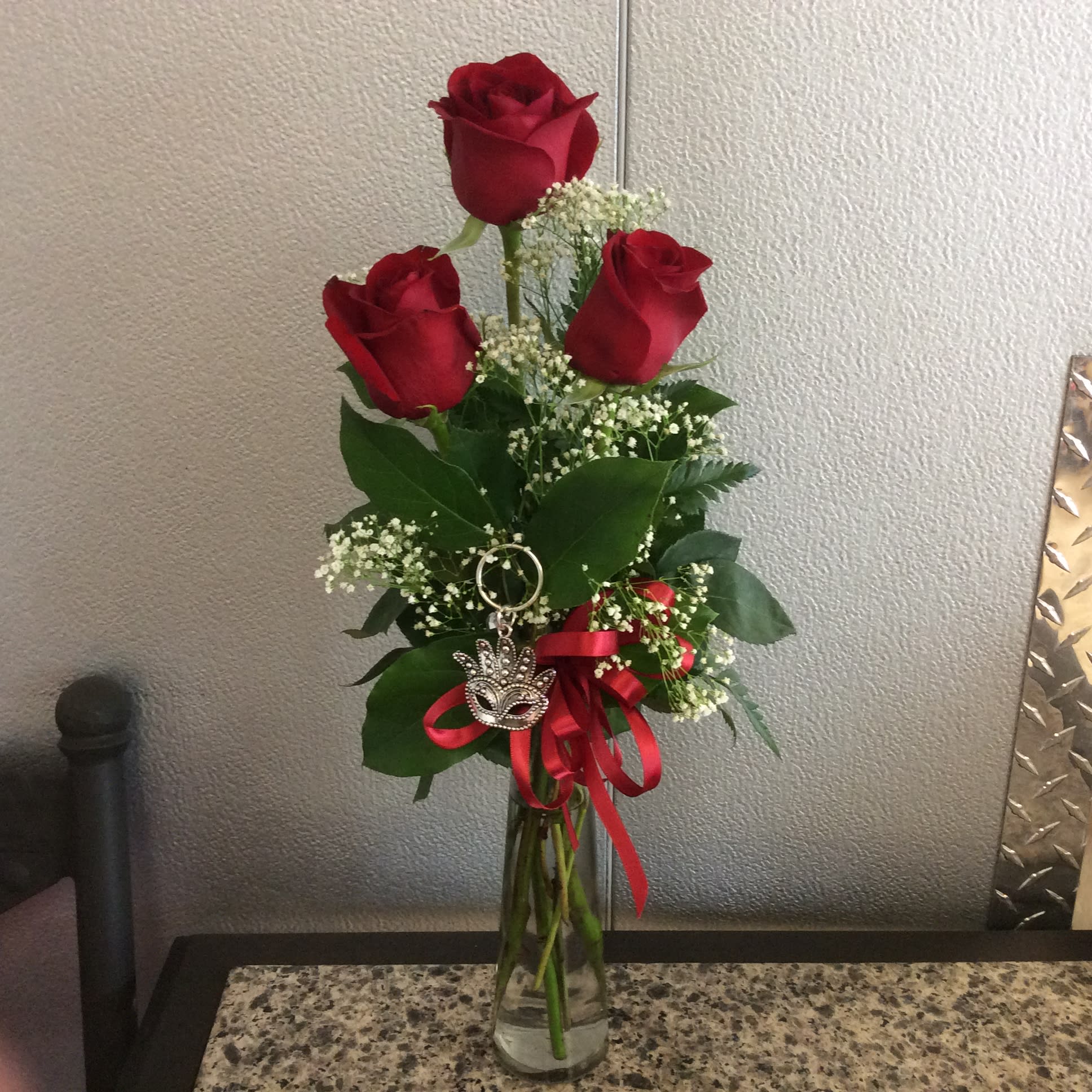 Three red roses in a clear glass vase with a red ribbon and charm