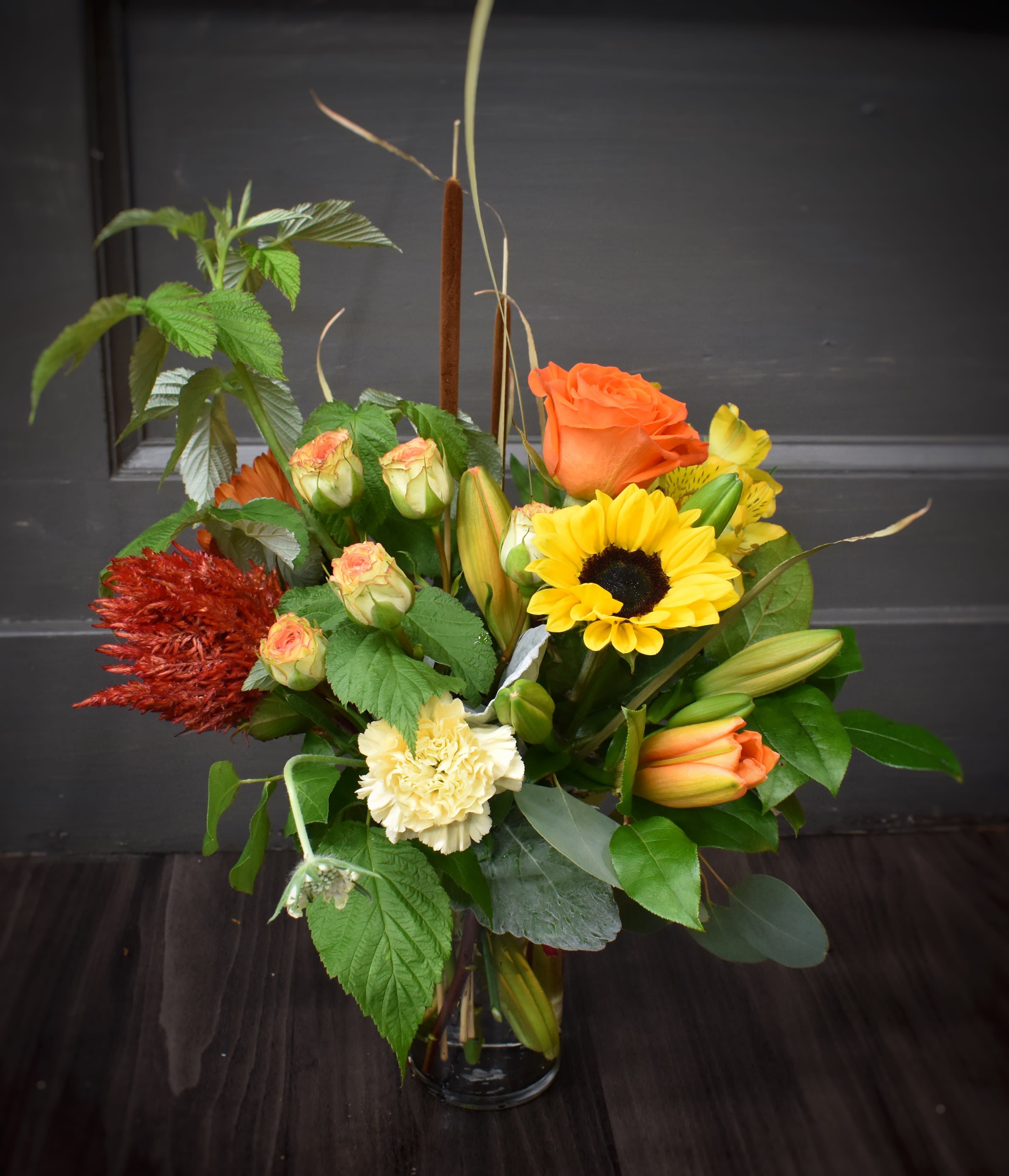 Mixed bouquet with orange roses, yellow sunflower, and lilies in a glass vase