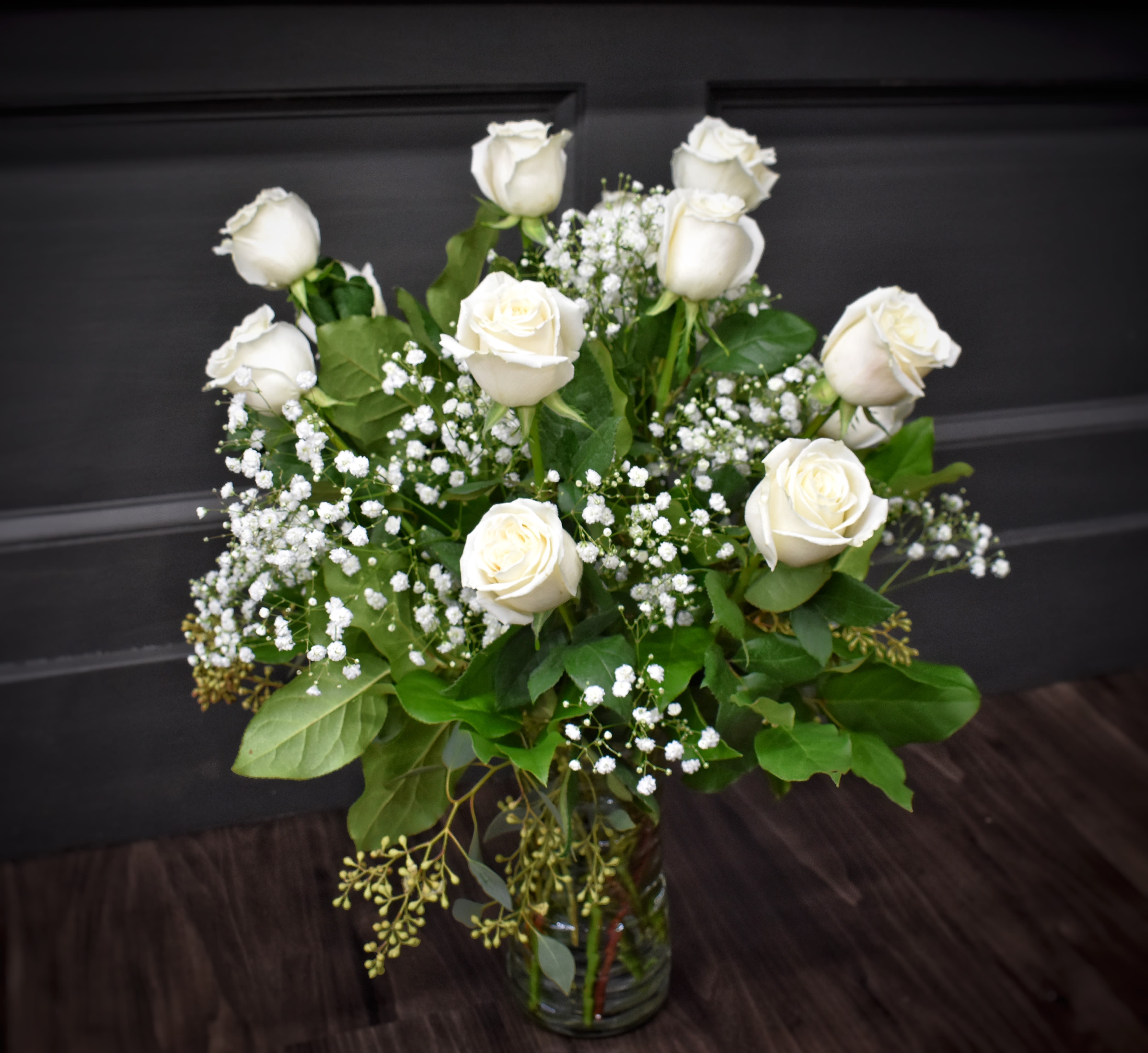 Tall arrangement of white roses and small white filler flowers in a clear glass vase.