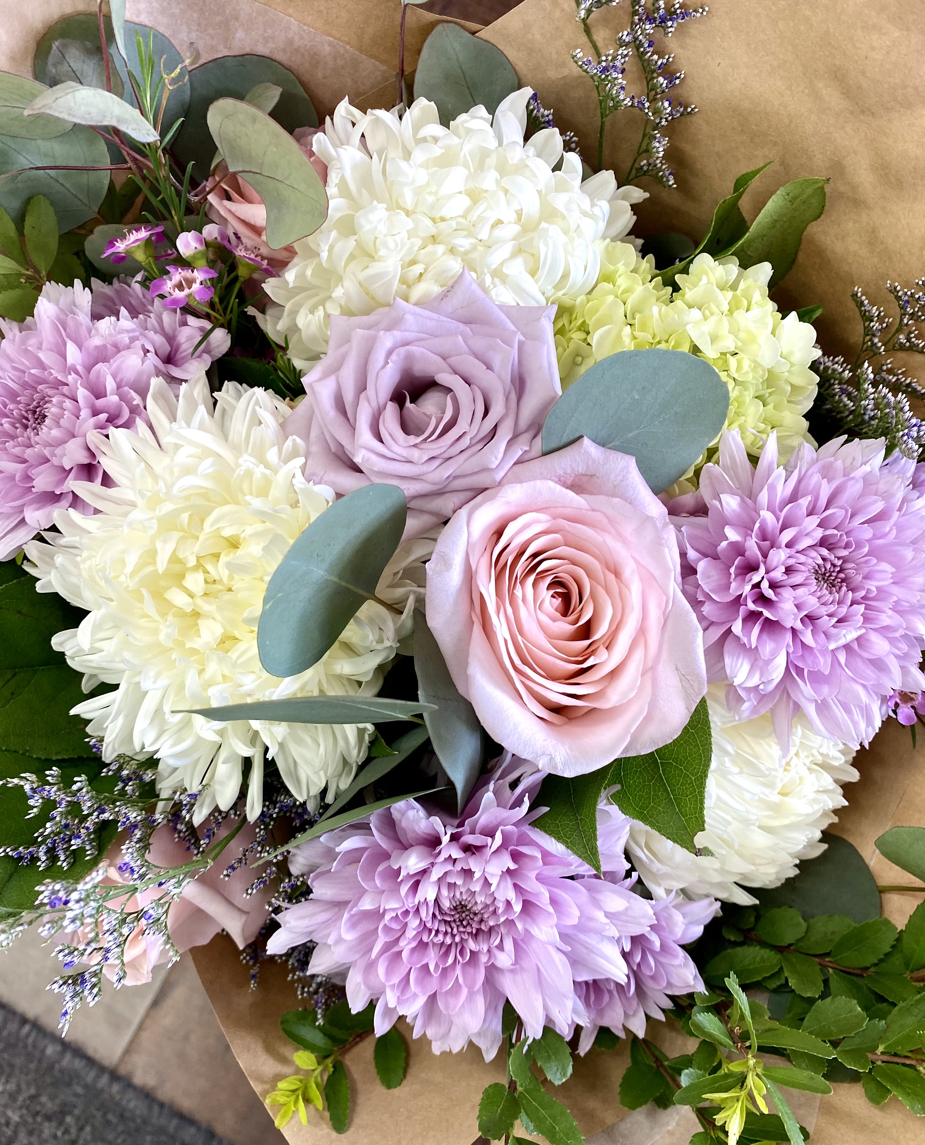 Hand-tied bouquet of pastel pink and lavender roses with white and lavender chrysanthemums in kraft paper.