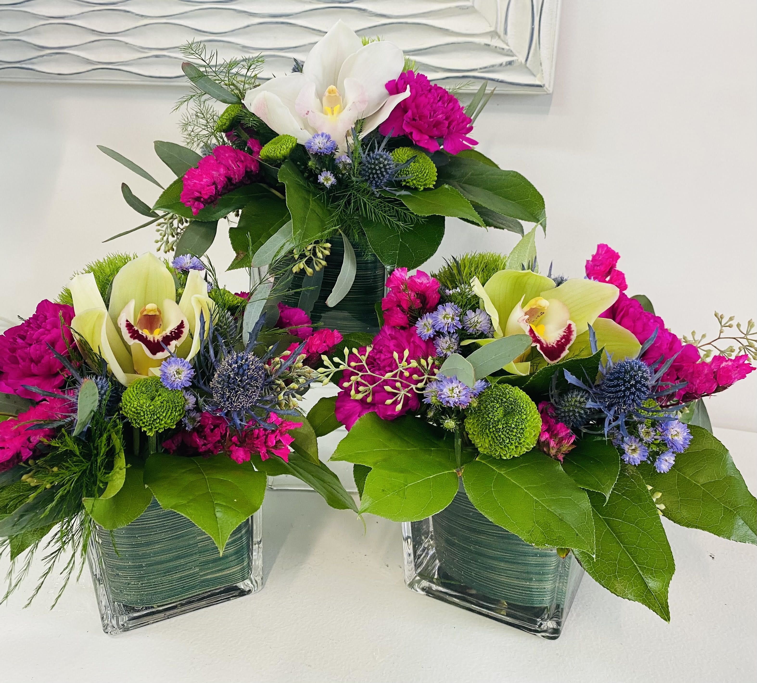 Three floral arrangements in square glass vases with orchids and bright pink blooms