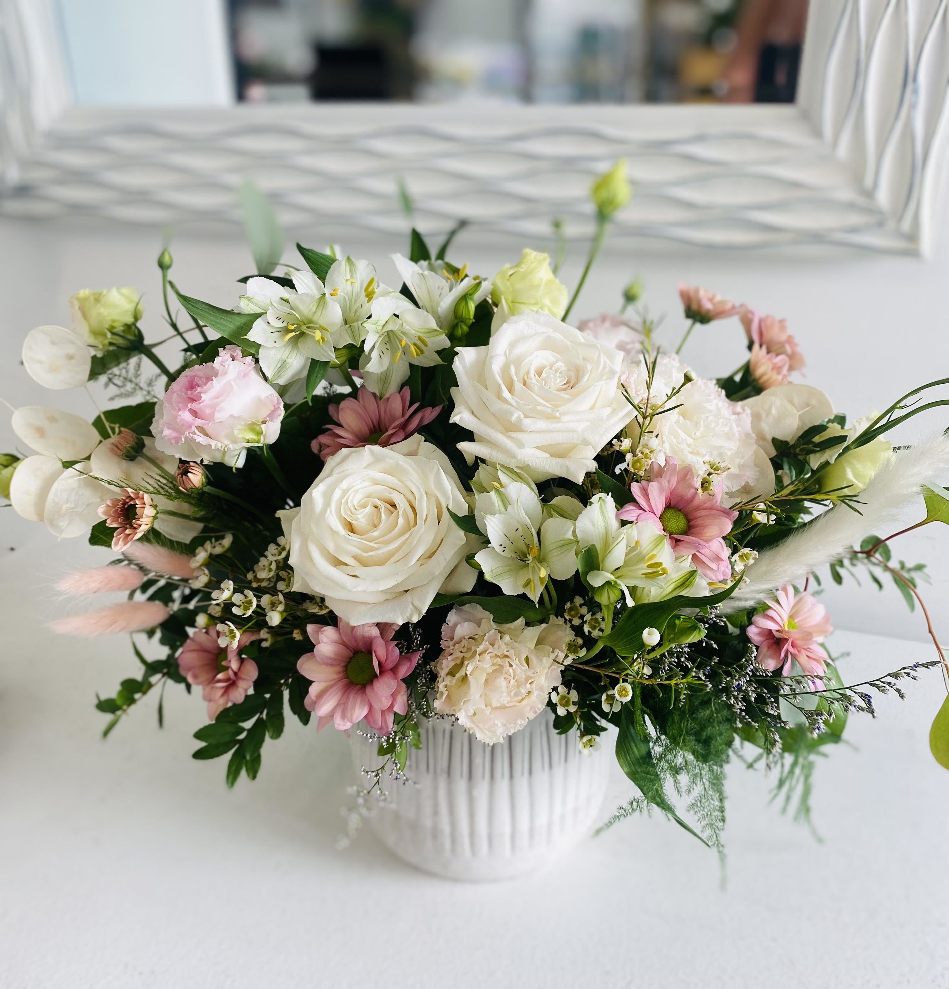 Low white ceramic vase filled with white roses, pink daisies, and soft pastel mixed flowers.