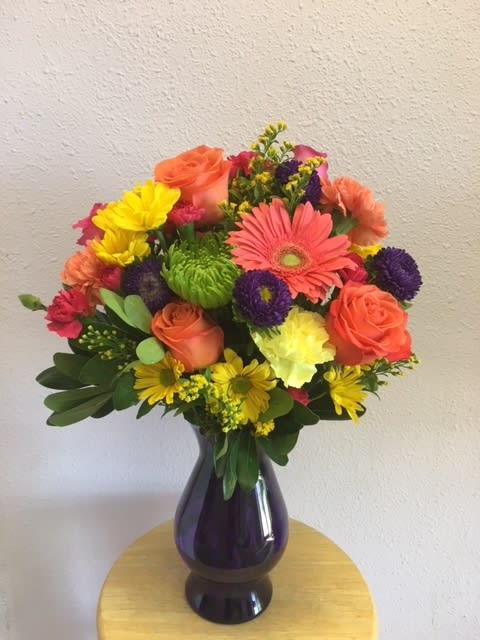 Magic Rainbow by Forever Flowers - Orange Roses, Purple Matsumoto Asters, Peach Gerbera Daisies, Green Fujies, Yellow Carnations, Yellow Daisies, Hot Pink Mini Carnations in a dark purple vase. 