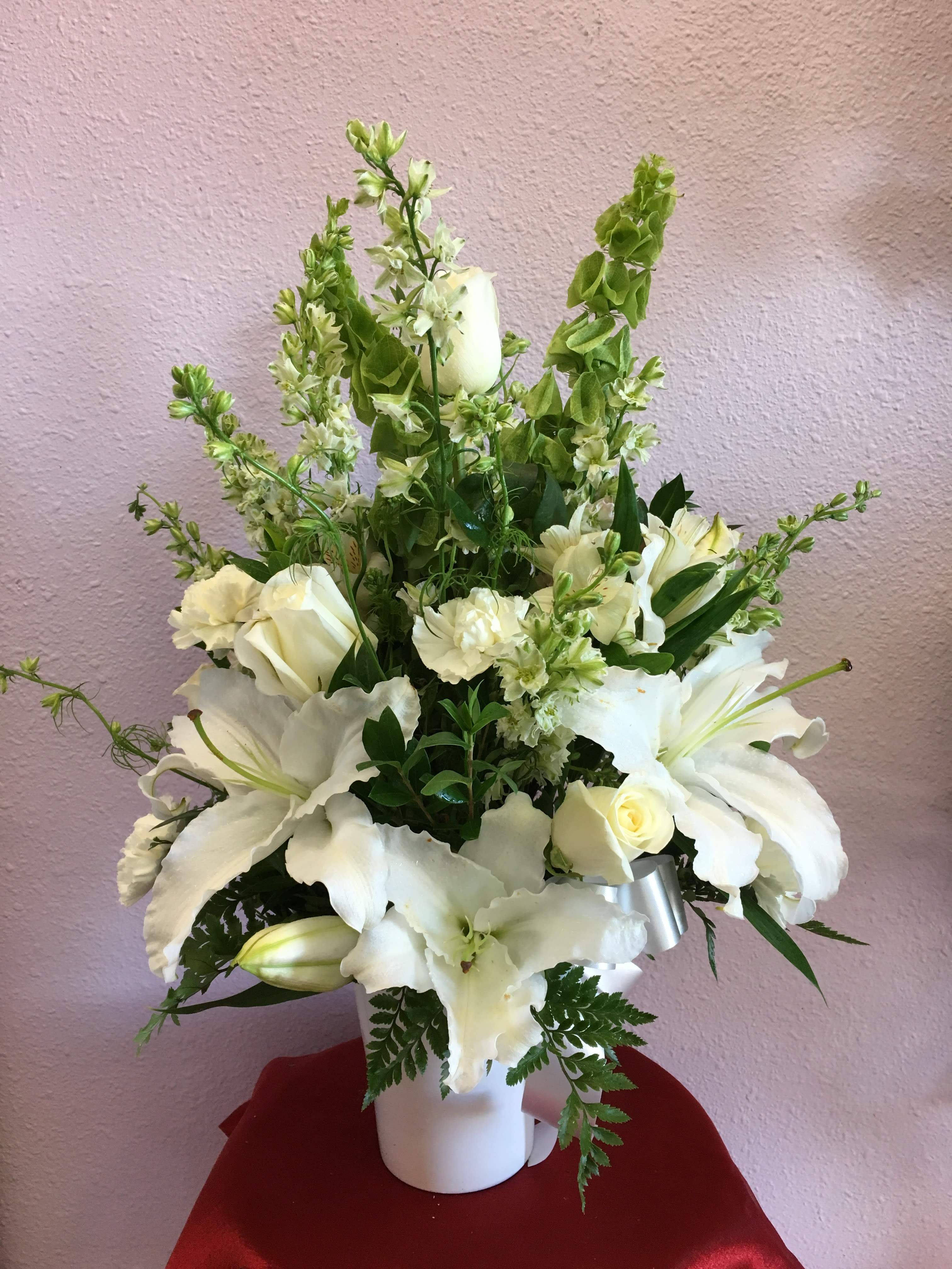 With Sympathy By Forever Flowers - Beautiful white arrangement with white lilies, white roses, bells or Ireland, white carnations, white larkspur and white alstroemerias in a white container.   