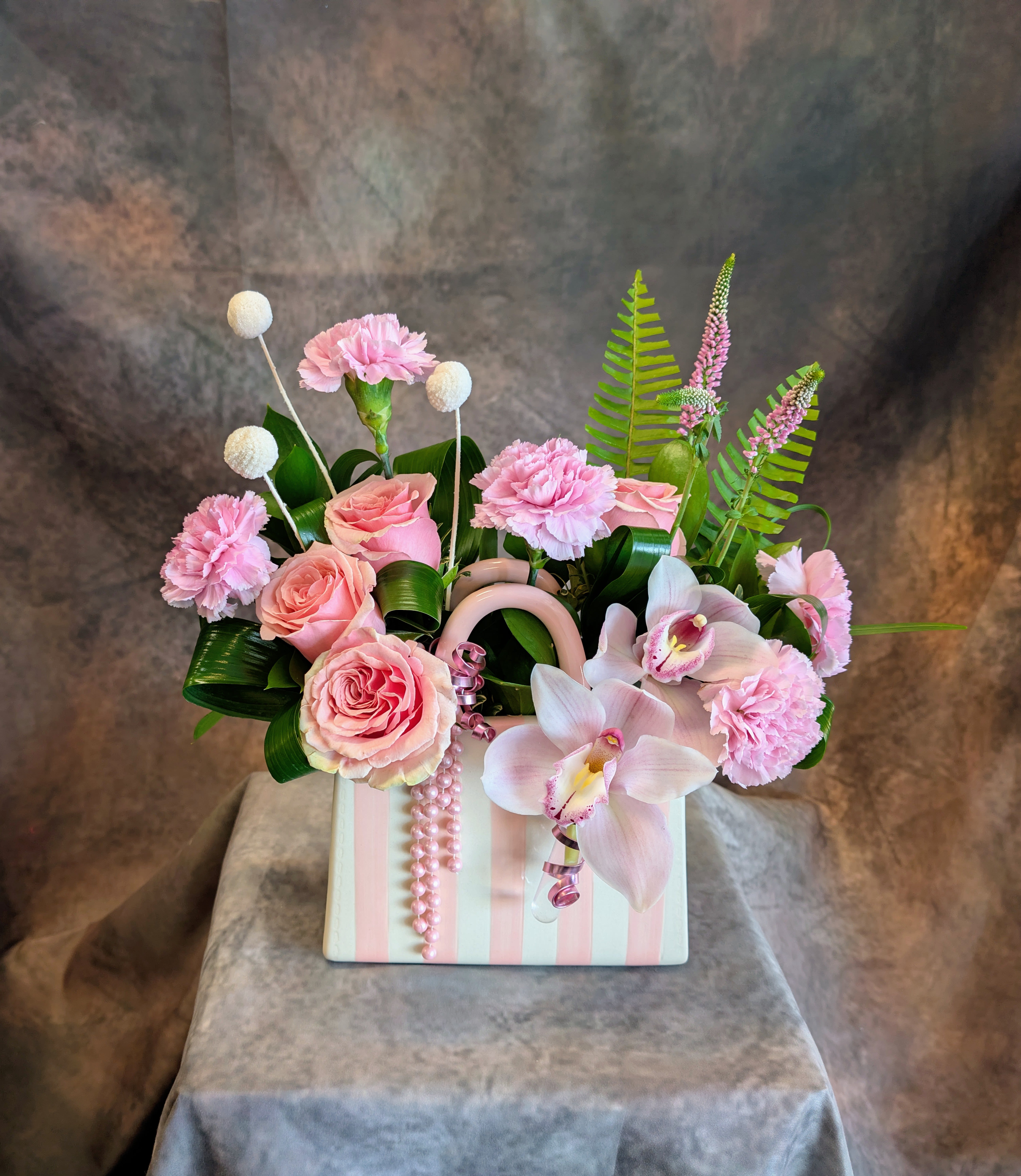 Miranda - Soft, feminine, and undeniably enchanting, Miranda is a romantic floral design handcrafted by Windmill Floral Expressions in Las Vegas. Designed in a chic blush pink striped ceramic handbag-inspired container with delicate pearl accents, this arrangement feels like a couture bouquet reimagined as floral art. Featuring elegant blush roses, soft pink carnations, graceful pink cymbidium orchids, textured billy balls, whimsical veronica, and sculptural tropical foliage, Miranda blends modern luxury with playful sophistication. The layered shades of pink create a dreamy, garden-inspired palette while the orchids add an elevated touch of refinement and movement. Curled greenery and delicate accents give the design a fashion-forward aesthetic that feels both whimsical and upscale — perfect for birthdays, Mother’s Day, baby celebrations, thank you gifts, anniversaries, or simply sending beauty “just because. Available for same-day flower delivery in Las Vegas, Henderson, Green Valley, Anthem, MacDonald Ranch, Inspirada, and surrounding areas. This one-of-a-kind floral handbag arrangement is designed to leave a lasting impression with its soft elegance, modern garden styling, and couture-inspired charm.