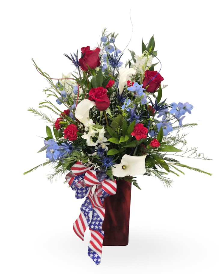 Patriotic Pride - Bold red roses and sculptural white calla lilies rise above spiky light-blue delphinium and fine-textured greenery in an airy, upright patriotic flower arrangement-perfect for a Fourth of July centerpiece or a Memorial Day tribute. The burgundy vase and a large red-white-and-blue ribbon with star-and-stripe detail give the design a celebratory, formal feel that also works for congratulations or to honor someone special.  This mixed-color arrangement includes roses, calla lilies, delphinium, carnations and other seasonal blooms arranged for height and layered texture. Finished in a tall burgundy vase with patriotic ribbon, the approximate dimensions are 25" x 16" x 10". Care: keep water fresh and trim stems every few days to maintain its upright, vibrant display.