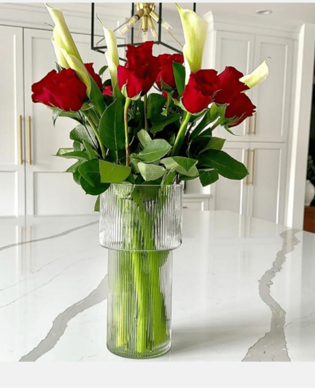 Red Rose and Calla Lily Arrangement in Clear Ribbed Vase - Tall white calla lilies rising above deep red roses create a striking vertical silhouette in this flower arrangement-perfect as an anniversary bouquet, Valentine's Day gift, or a refined sympathy tribute. The red-and-white contrast reads as timelessly romantic and polished, the kind of presentation that makes the person you're thinking of feel truly honored and seen.  Compact yet upright, the design places glossy green foliage around the middle and base for a clean, modern profile. Flowers are delivered in a clear ribbed glass vase that shows the long stems and adds subtle texture while keeping the look contemporary. The arrangement's height and structured lines make it ideal for tabletops, entry consoles, or beside a mantle; order with confidence for any occasion that calls for elegant, heartfelt expression.