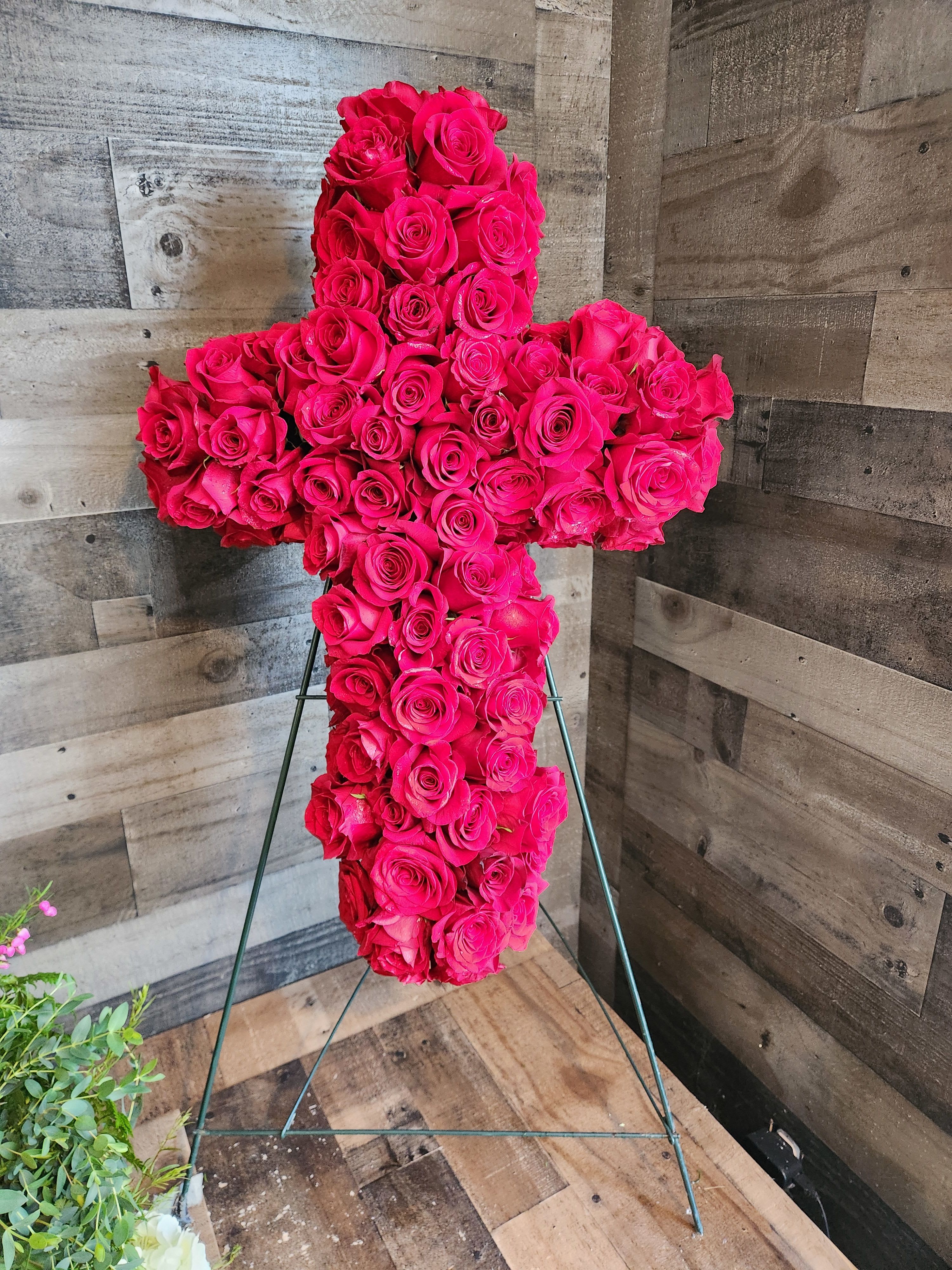 Standing cross of hot pink roses on a metal easel against a wood wall.