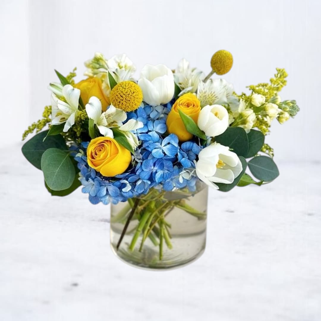 Bouquet of yellow roses, blue hydrangeas, and white tulips in a glass vase