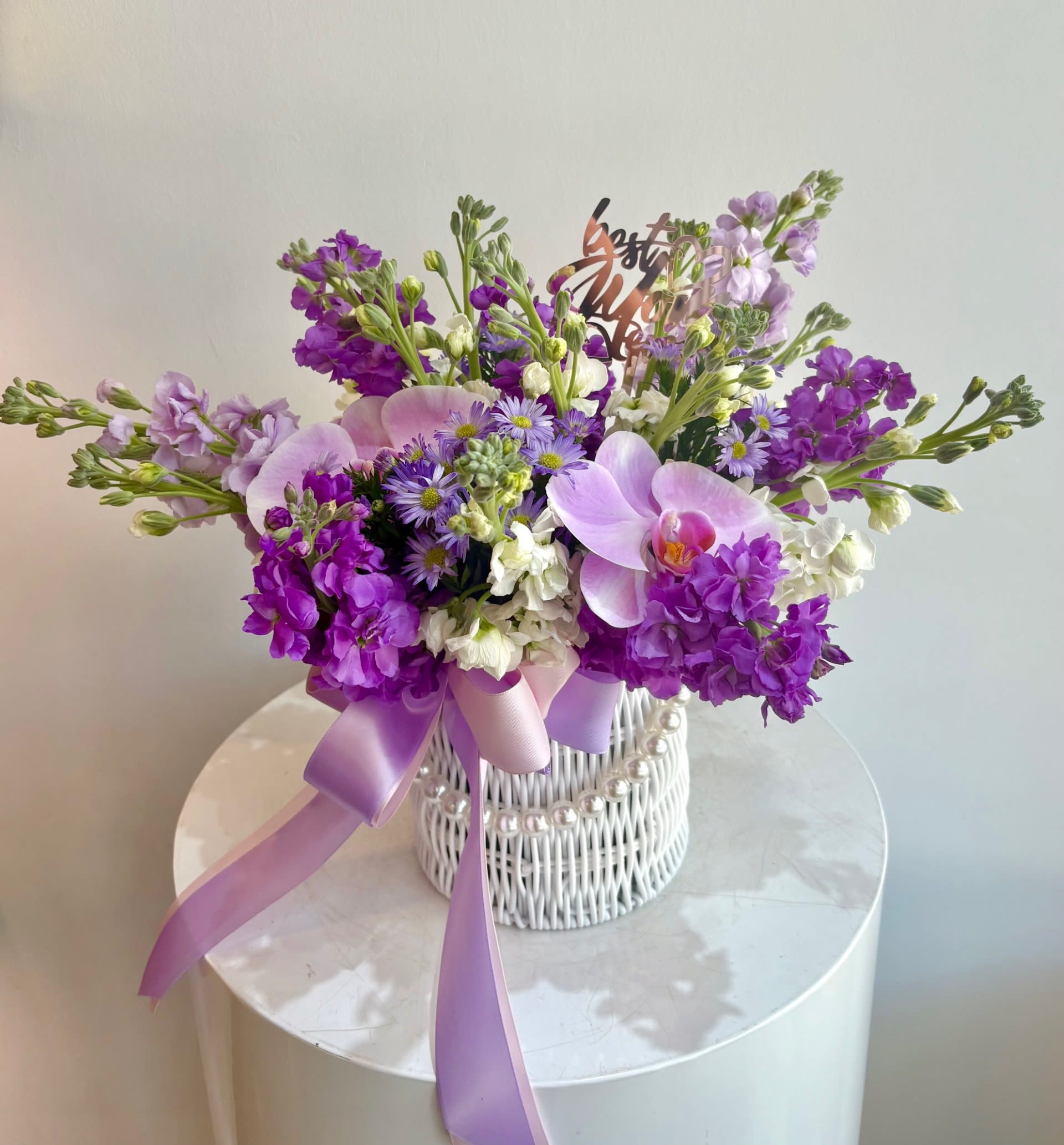 Purple and white floral arrangement in a white wicker basket with a lavender ribbon