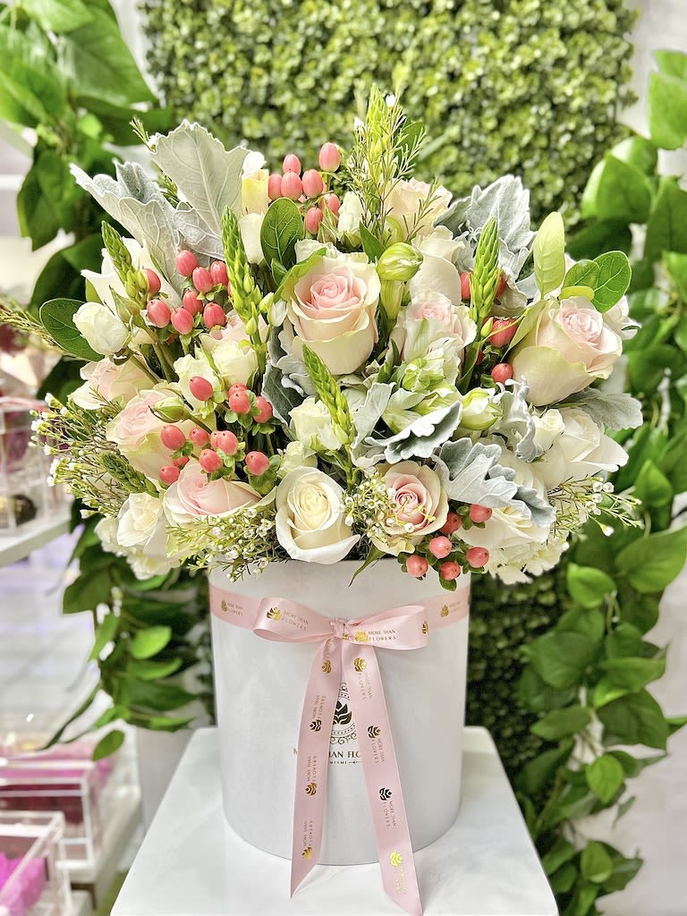 Round hatbox arrangement of pale pink and white roses with pink berries and foliage, tied with a pink ribbon.