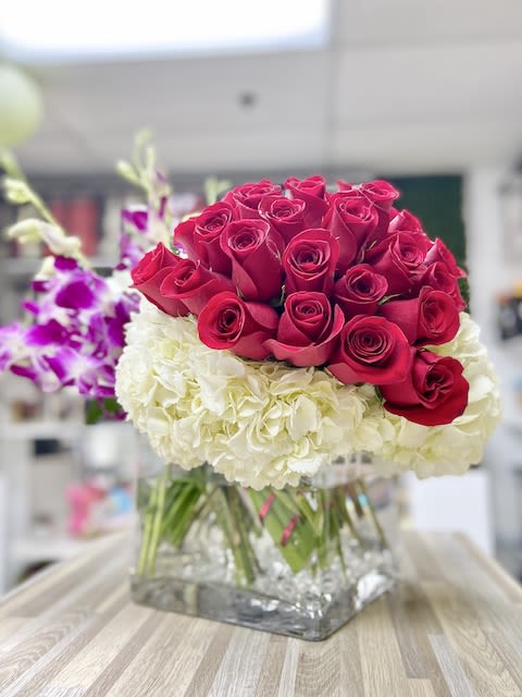Red roses arranged over white hydrangeas with purple orchids in a clear square glass vase