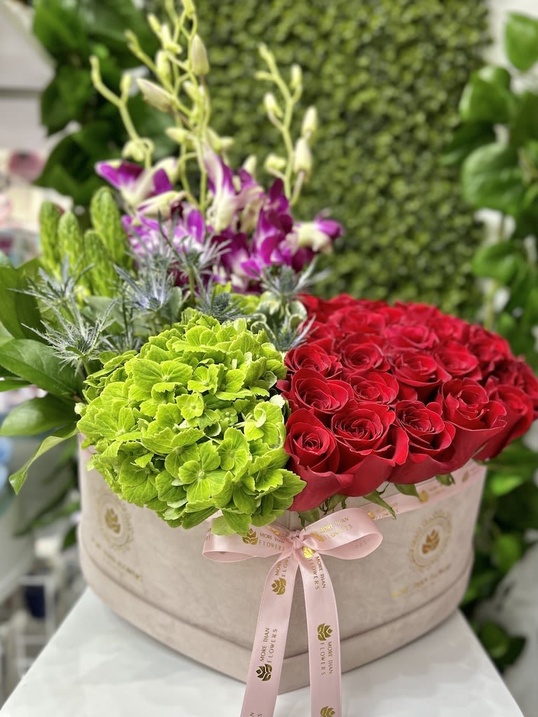 Round hatbox arrangement with red roses, green hydrangeas, and purple orchids tied with a pink ribbon.