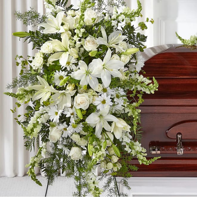 White funeral spray with lilies, roses, and daisies beside a casket