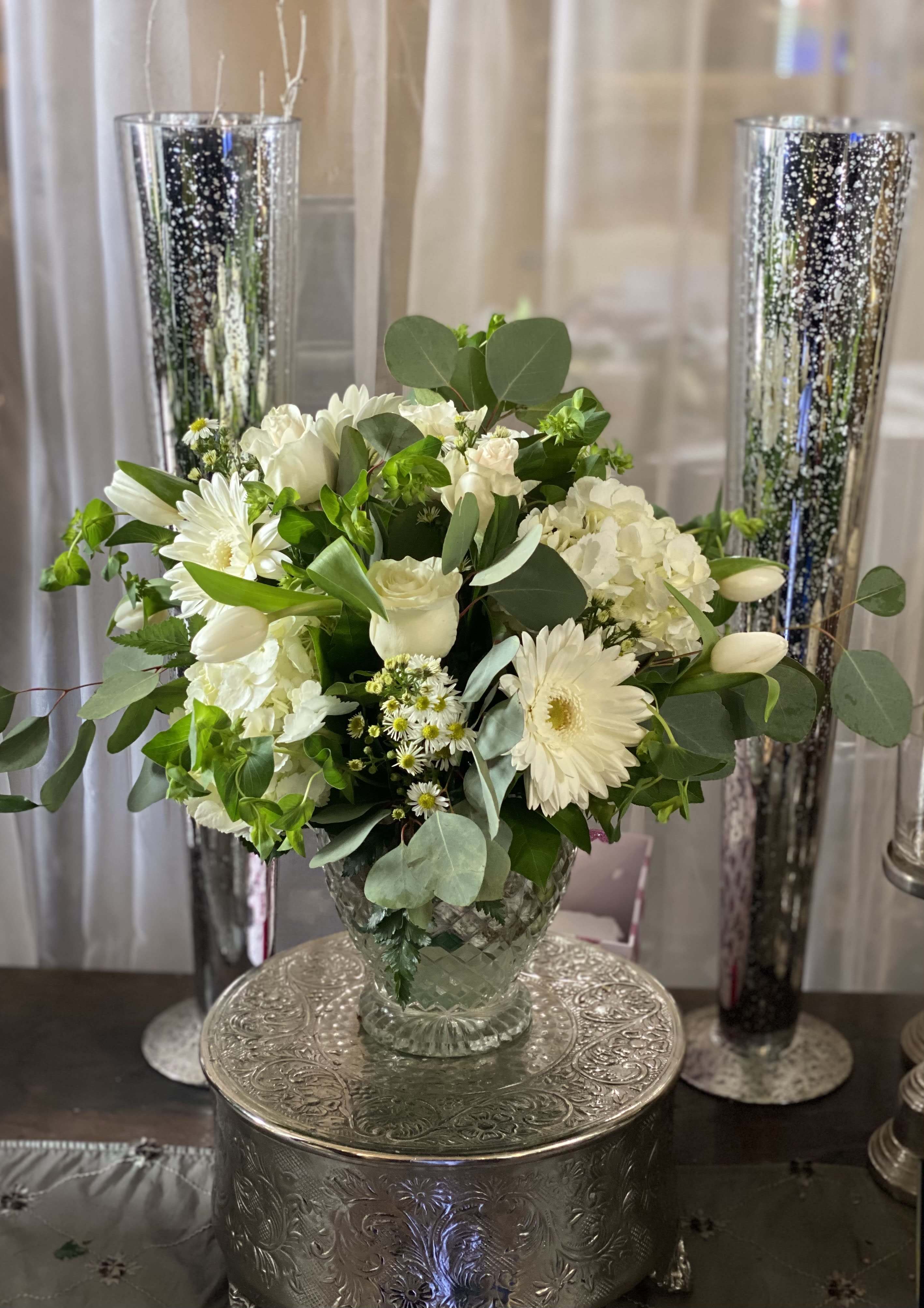 White floral arrangement in a clear glass vase with tall silver candle holders behind it