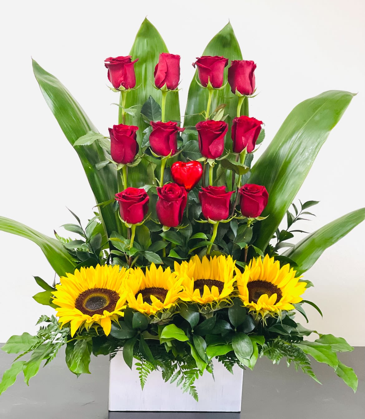 Red roses and sunflowers arranged in a white box with large green leaves