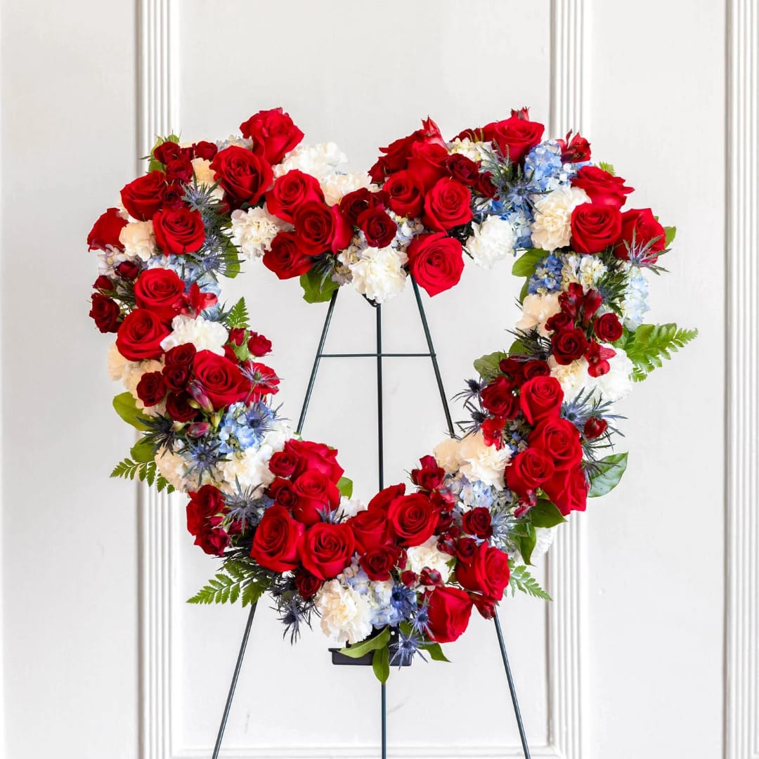 Heart-shaped floral wreath of red roses, white flowers, and blue accents on a stand