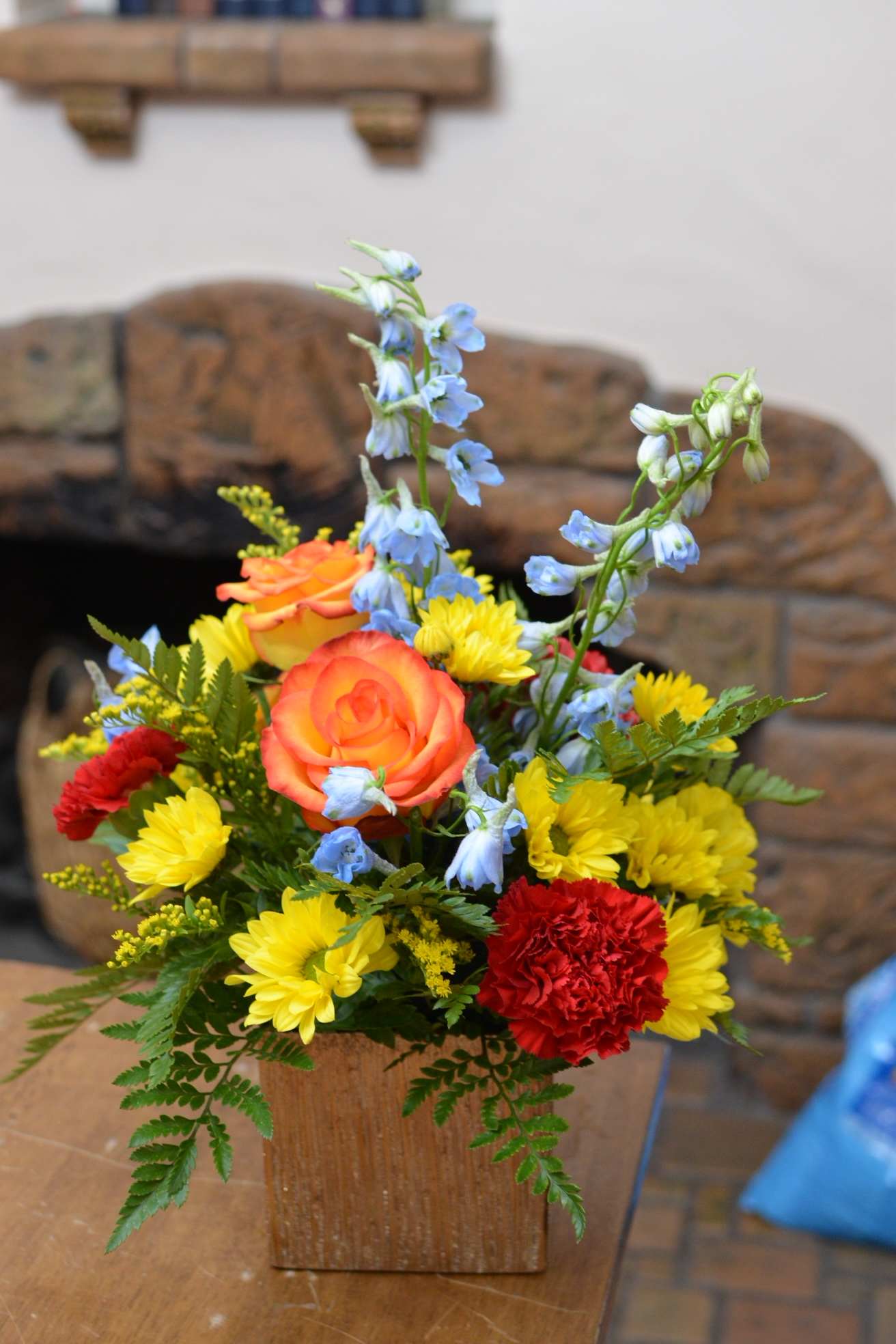 Colorful flower arrangement in a wooden cube vase with roses, daisies, and blue delphinium