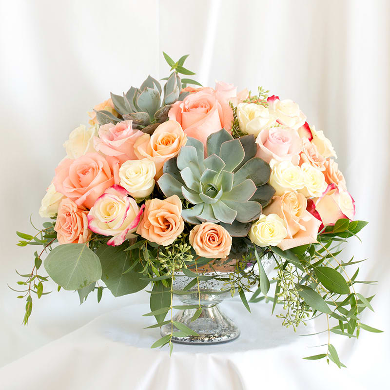 Peach and cream roses arranged with succulents in a glass pedestal vase