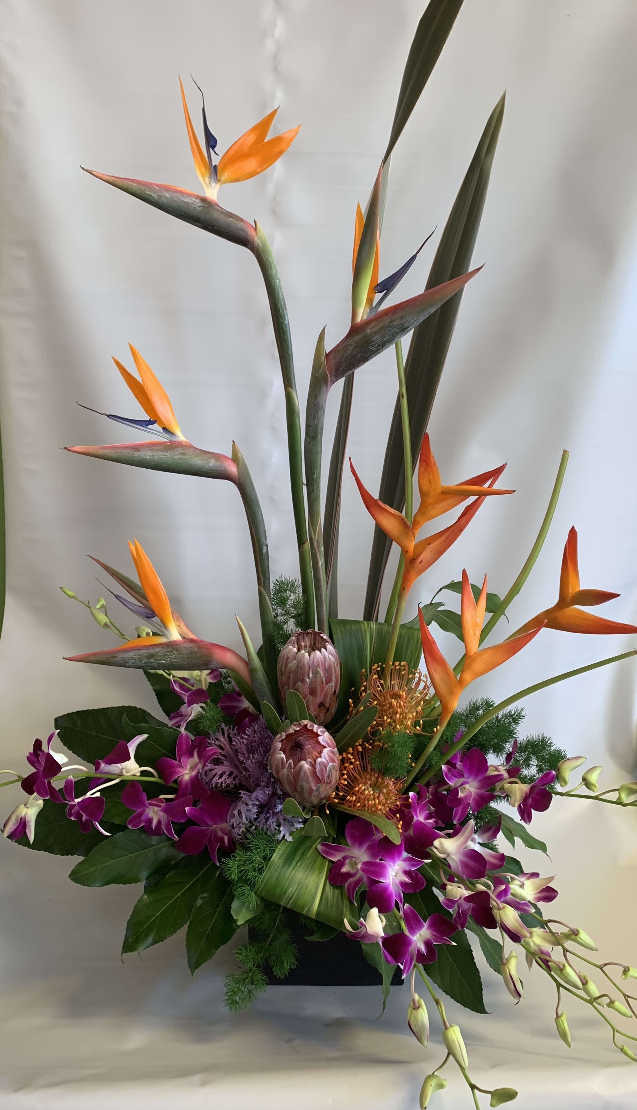 Tall tropical arrangement with orange bird-of-paradise and purple orchids in a black container