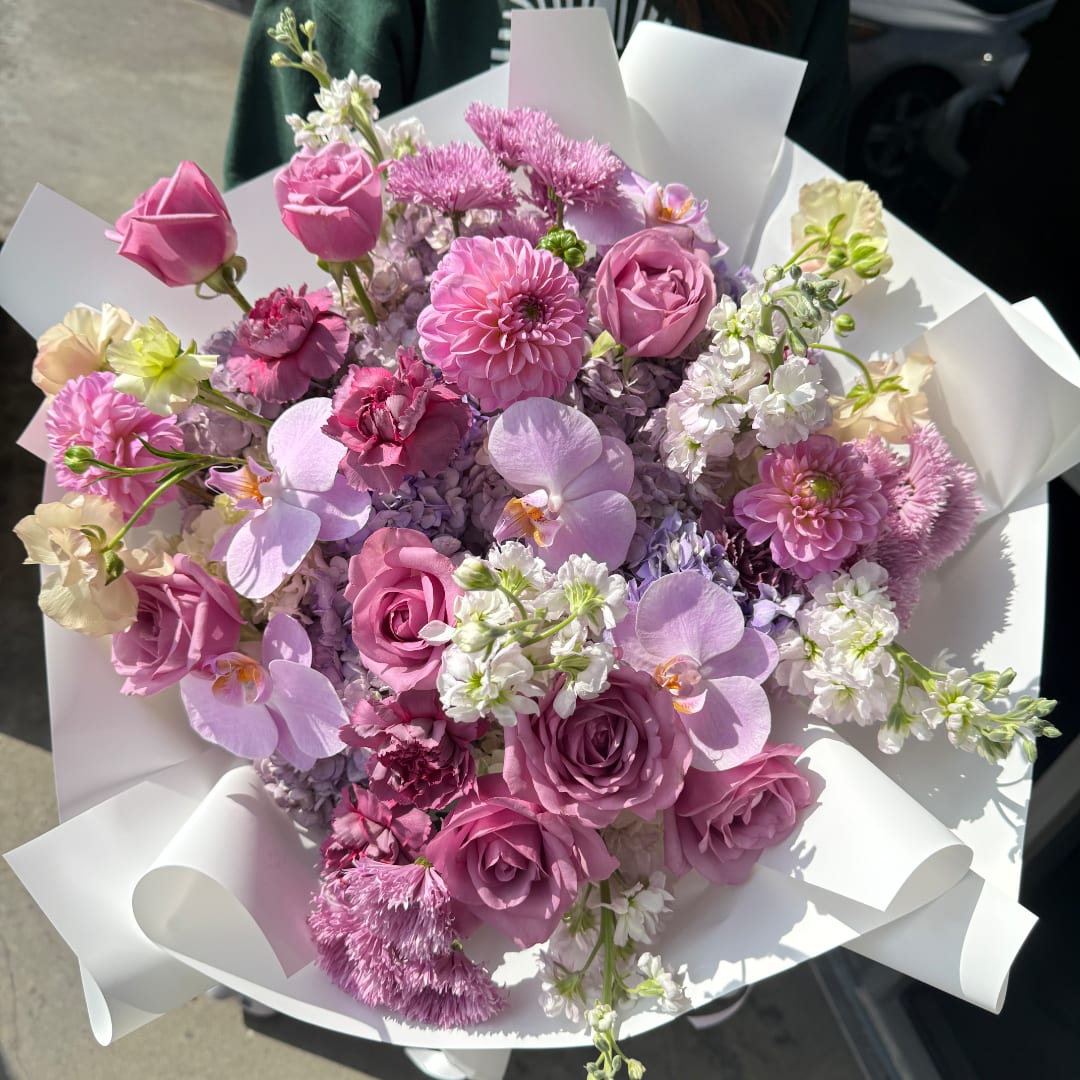 Pink and lavender bouquet with roses, orchids, and mixed blooms wrapped in white paper