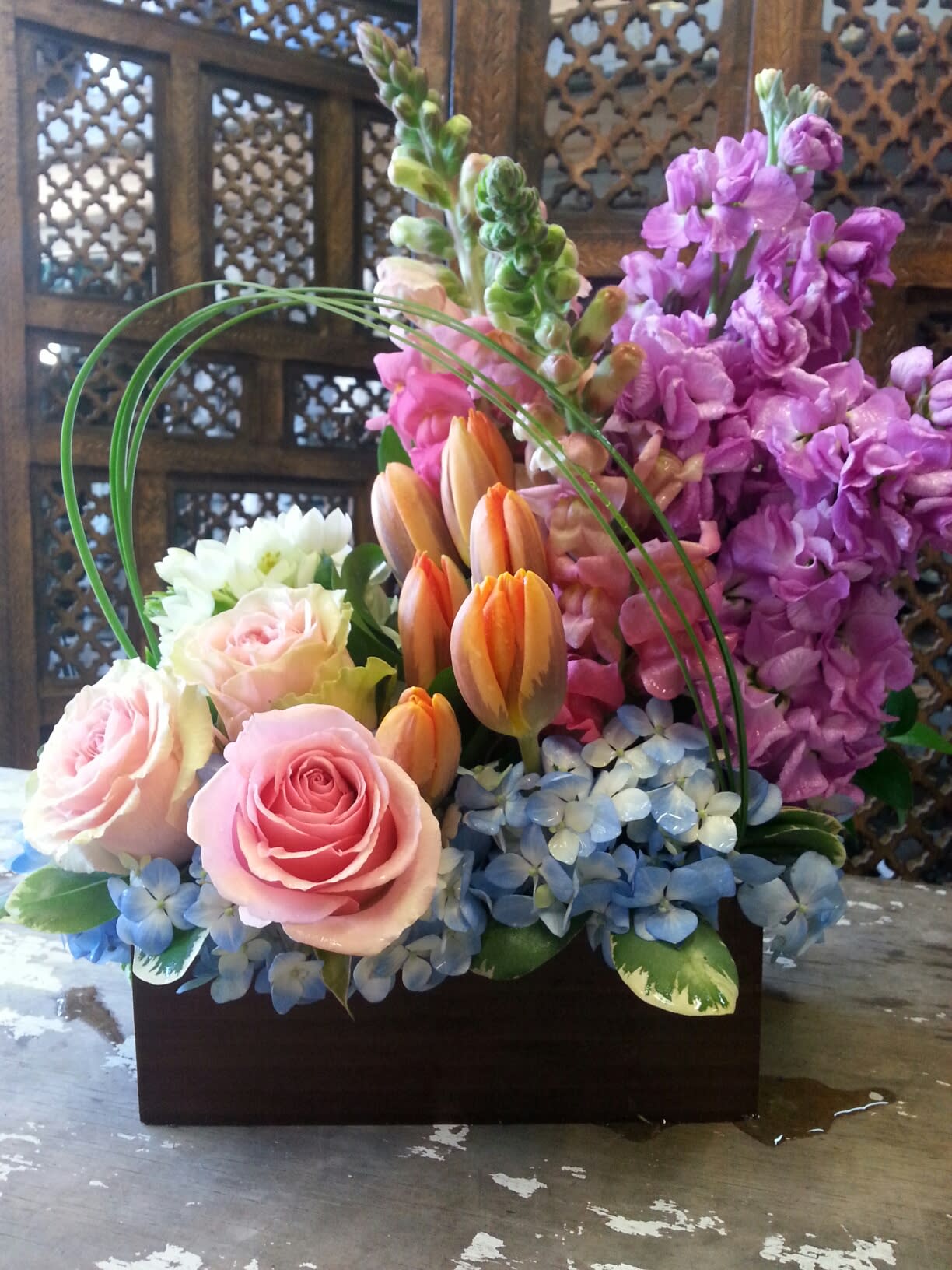 Mixed bouquet of pink roses, orange tulips, and purple blooms in a dark container