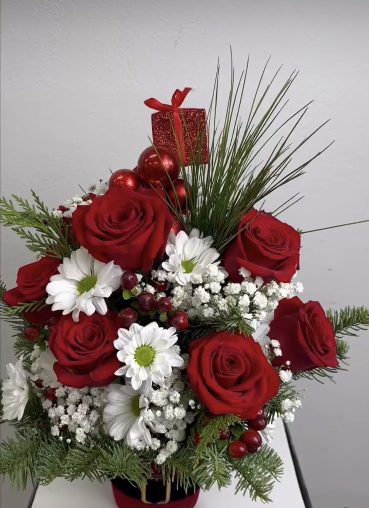 Red roses and white daisies in a festive holiday arrangement with a gift box topper
