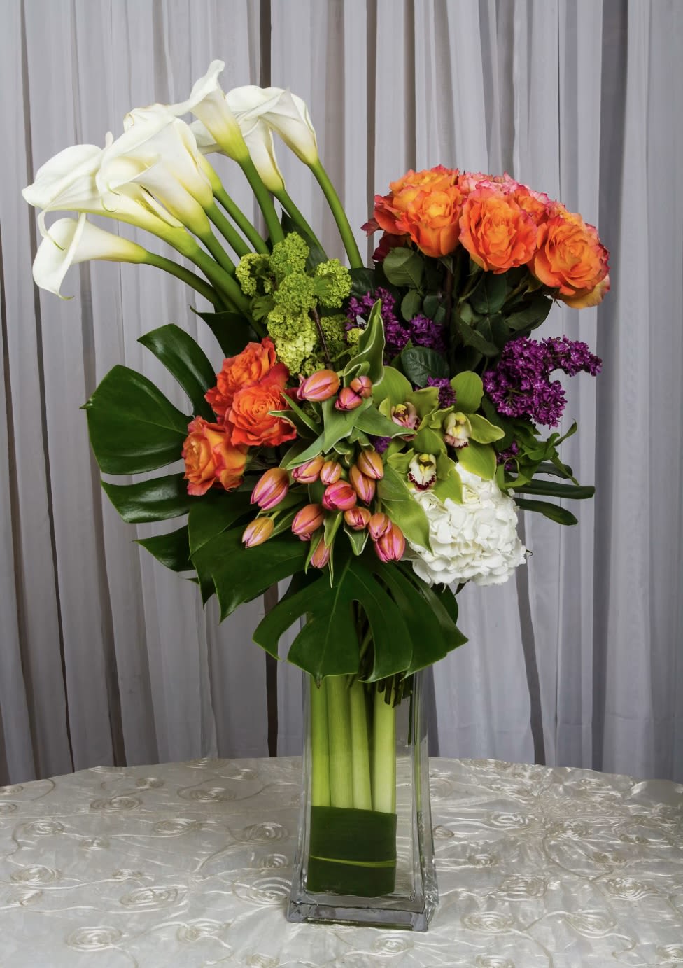 Tall bouquet of white calla lilies, orange roses, and purple flowers in a glass vase