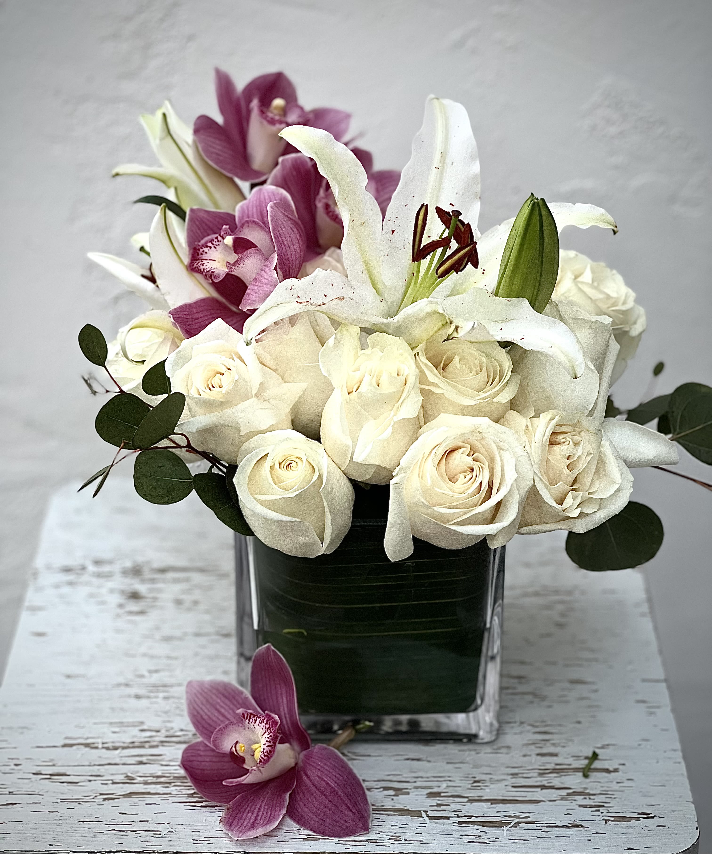 White roses and lilies arranged in a square glass vase with purple orchids