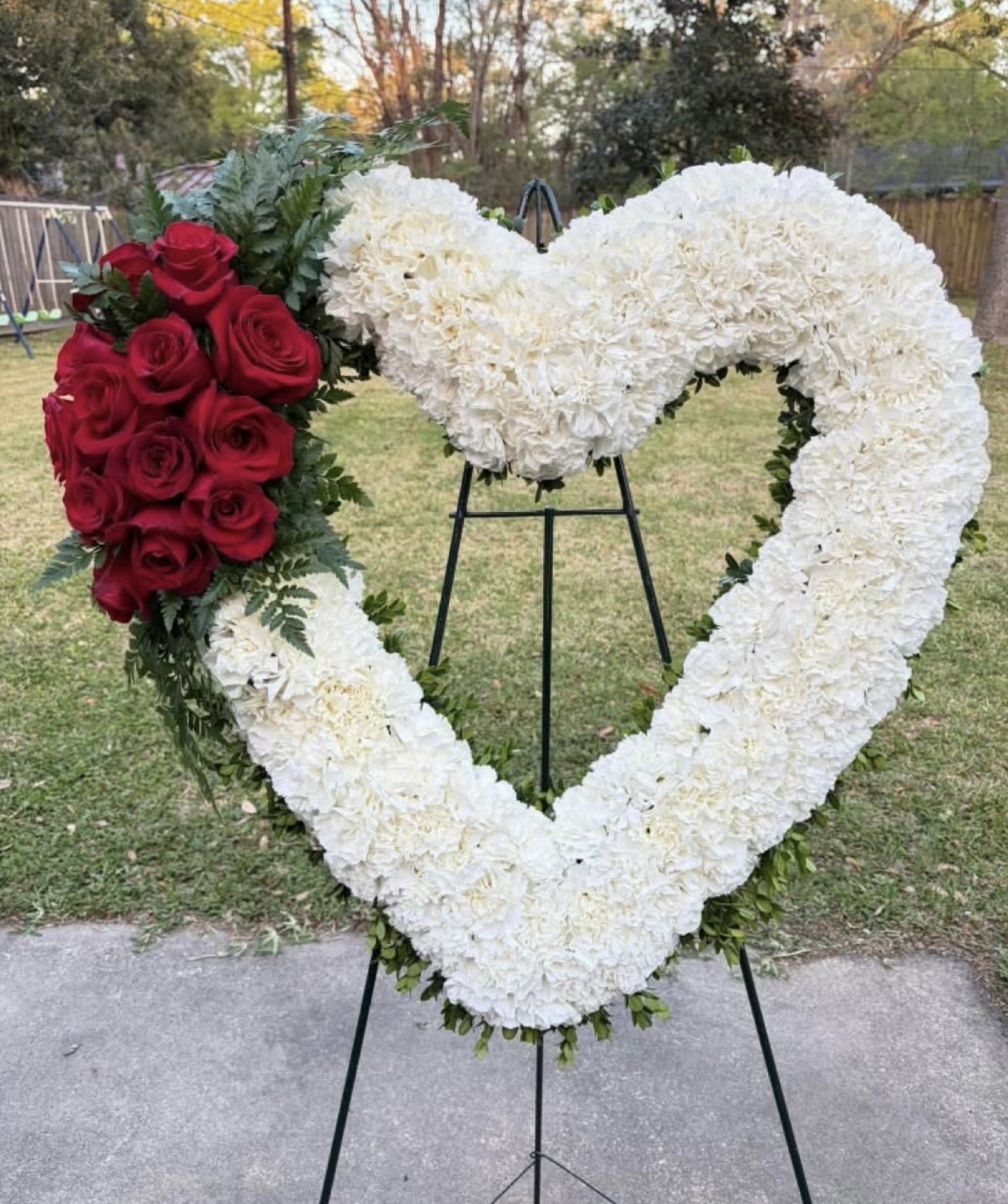 Eternal Love Heart Wreath - Honor a life beautifully lived with our Eternal Love Heart Wreath. This elegant heart-shaped arrangement features a lush base of soft white blooms, symbolizing peace and remembrance, accented with a striking cluster of deep red roses to represent enduring love and respect. Designed with care and artistry, this standing wreath offers a heartfelt tribute for funerals and memorial services.  Crafted fresh with premium flowers and presented on a sturdy easel, it creates a graceful and meaningful presence at any service.  Order now and let your condolences be expressed with timeless beauty and dignity.