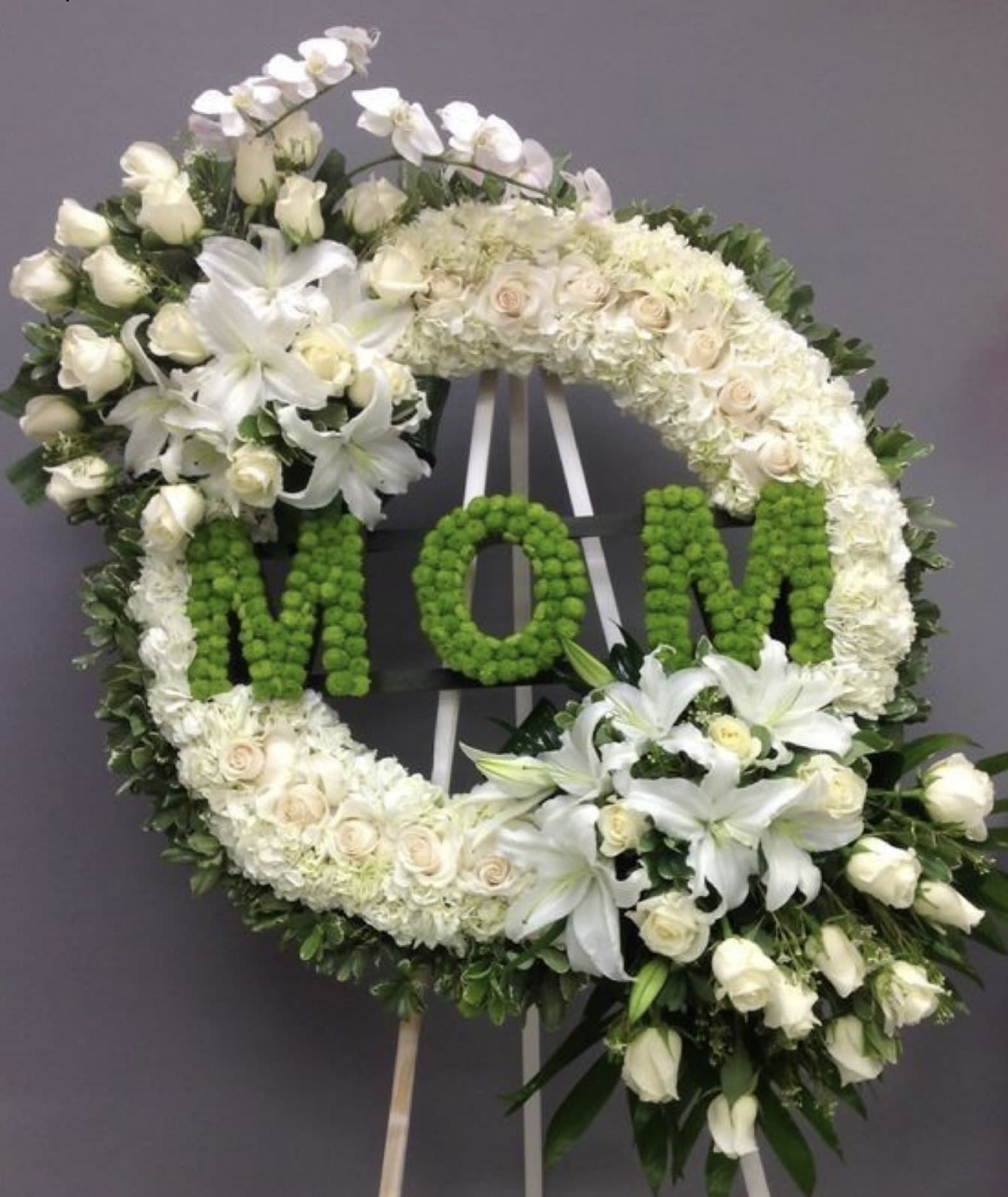 Heavenly Tribute “Mom” Wreath - Celebrate the life and love of a cherished mother with our Heavenly Tribute “Mom” Wreath. This elegant circular arrangement is crafted with soft white blooms symbolizing peace, purity, and remembrance, beautifully accented with delicate lilies and roses. The vibrant green “MOM” lettering adds a deeply personal and heartfelt touch, making it a meaningful tribute for funeral and memorial services.  Designed with premium fresh flowers and displayed on a standing easel, this wreath offers a graceful expression of love, honor, and eternal connection.  Order today to create a touching and unforgettable tribute.