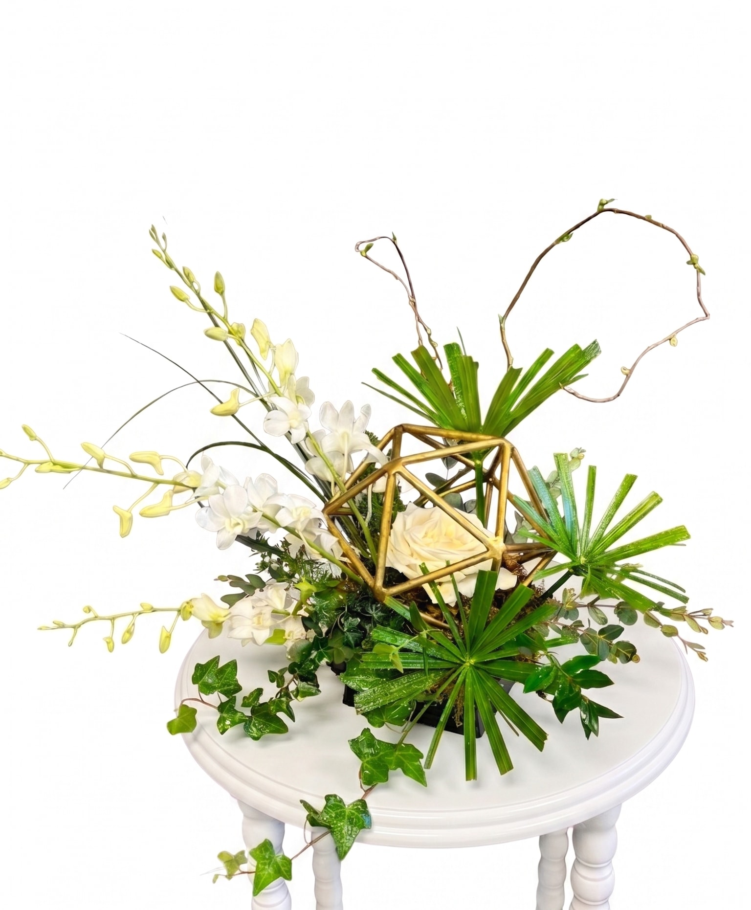 The Orchid Prism Bouquet - Make a bold statement with The Orchid Prism Bouquet, a captivating arrangement designed for those who appreciate cutting-edge, sculptural floral art. At the heart of this piece is a striking gold geometric prism that frames a single, pristine white rose. Dramatic, cascading stems of elegant white orchids break through the structure, perfectly balanced by the sharp lines of fresh fan palms, trailing ivy, and winding curly willow branches.  With its distinct architectural vibe, this arrangement brings a sophisticated, high-end gallery feel into any space.