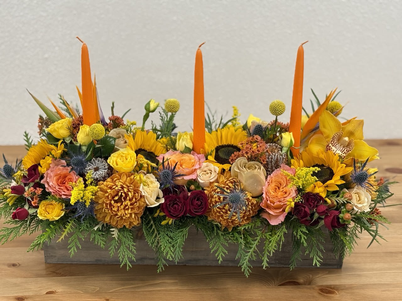 Long floral centerpiece with orange candles and mixed yellow, pink, and burgundy blooms