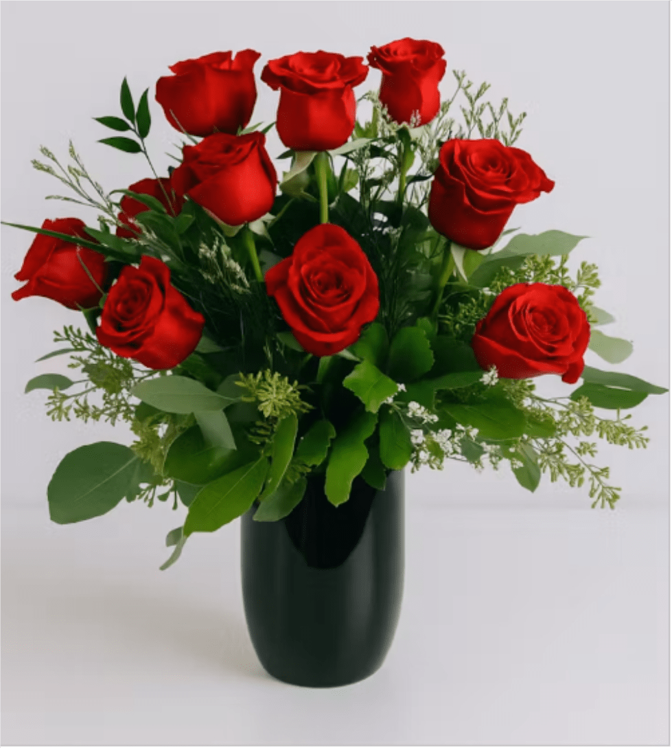 Red Rose Vase Arrangement - Red Roses in Dark Vase - Velvety red roses arranged in a compact, rounded bouquet make an immediate, romantic impression-perfect as an anniversary bouquet, Valentine's Day gift, or a simple "I'm thinking of you" surprise. The dozen red roses sit upright in a dark glass vase, the vivid crimson blooms contrasted against deep green foliage for a classic, timeless look that reads formal without feeling fussy.  Mixed airy filler and varied greenery soften the silhouette and add delicate texture around the full-petaled roses, while the dark cylindrical vase anchors the arrangement with modern elegance. Sized to sit comfortably on a table or desk, this arrangement arrives ready to display; it's the kind of floral gift that will make the person you care about feel truly seen and celebrated.