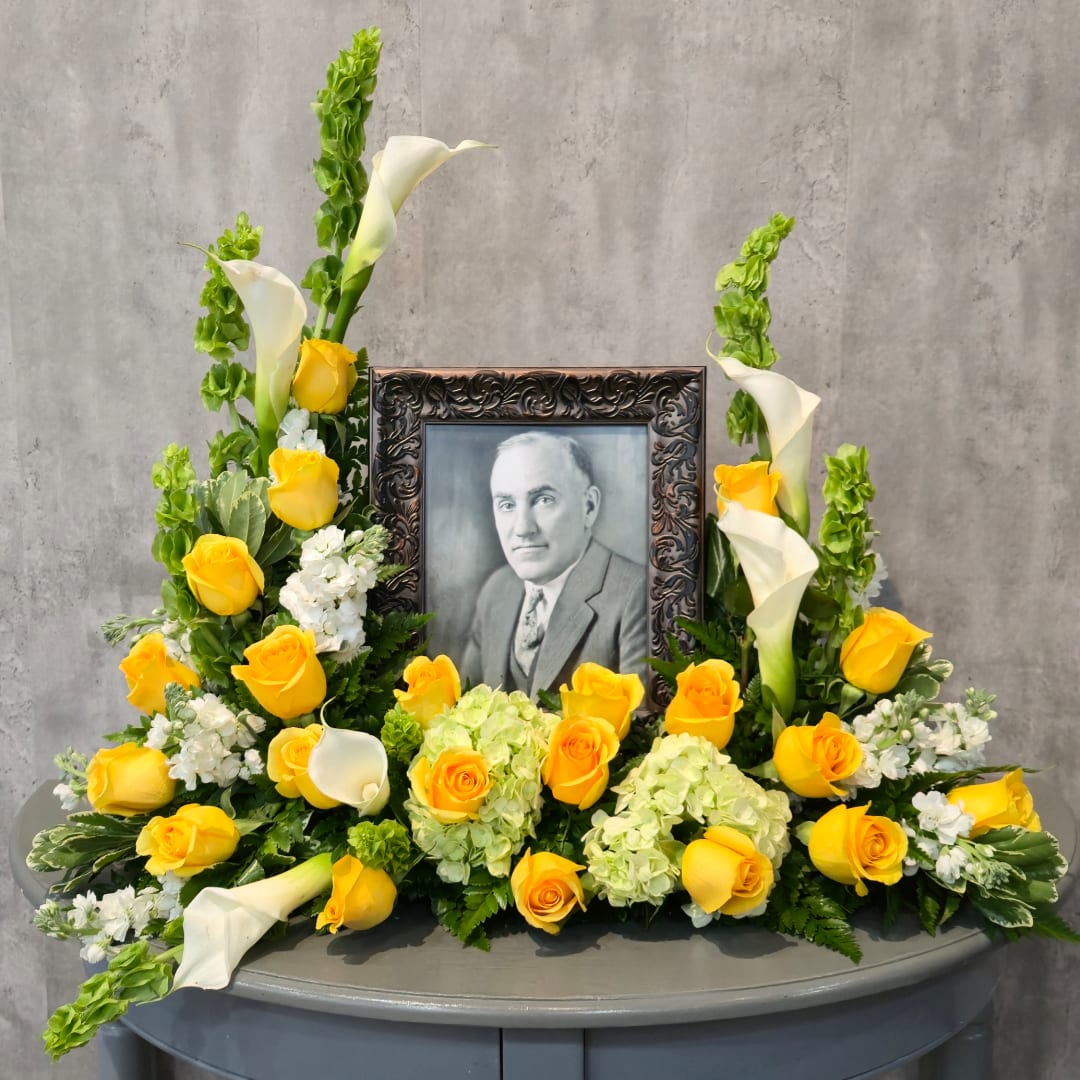 Yellow roses and white calla lilies arranged around a framed portrait